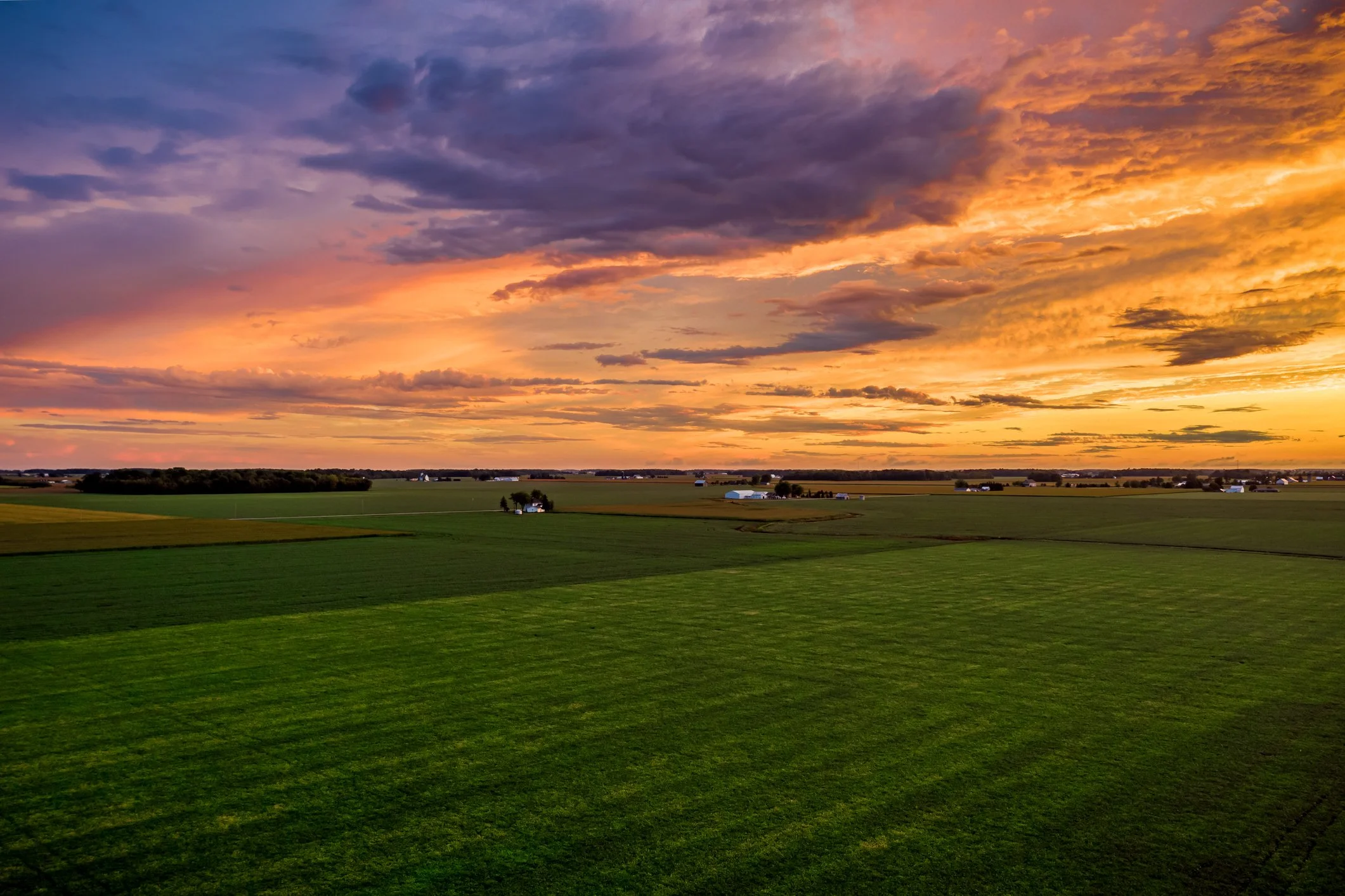 A vast rural landscape at sunset with green fields and scattered farm buildings under a colorful sky with clouds.