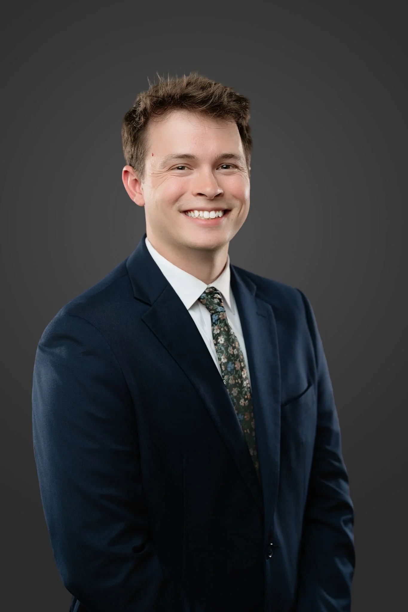 A young man with short brown hair in a suit, smiling with a dark gray background.