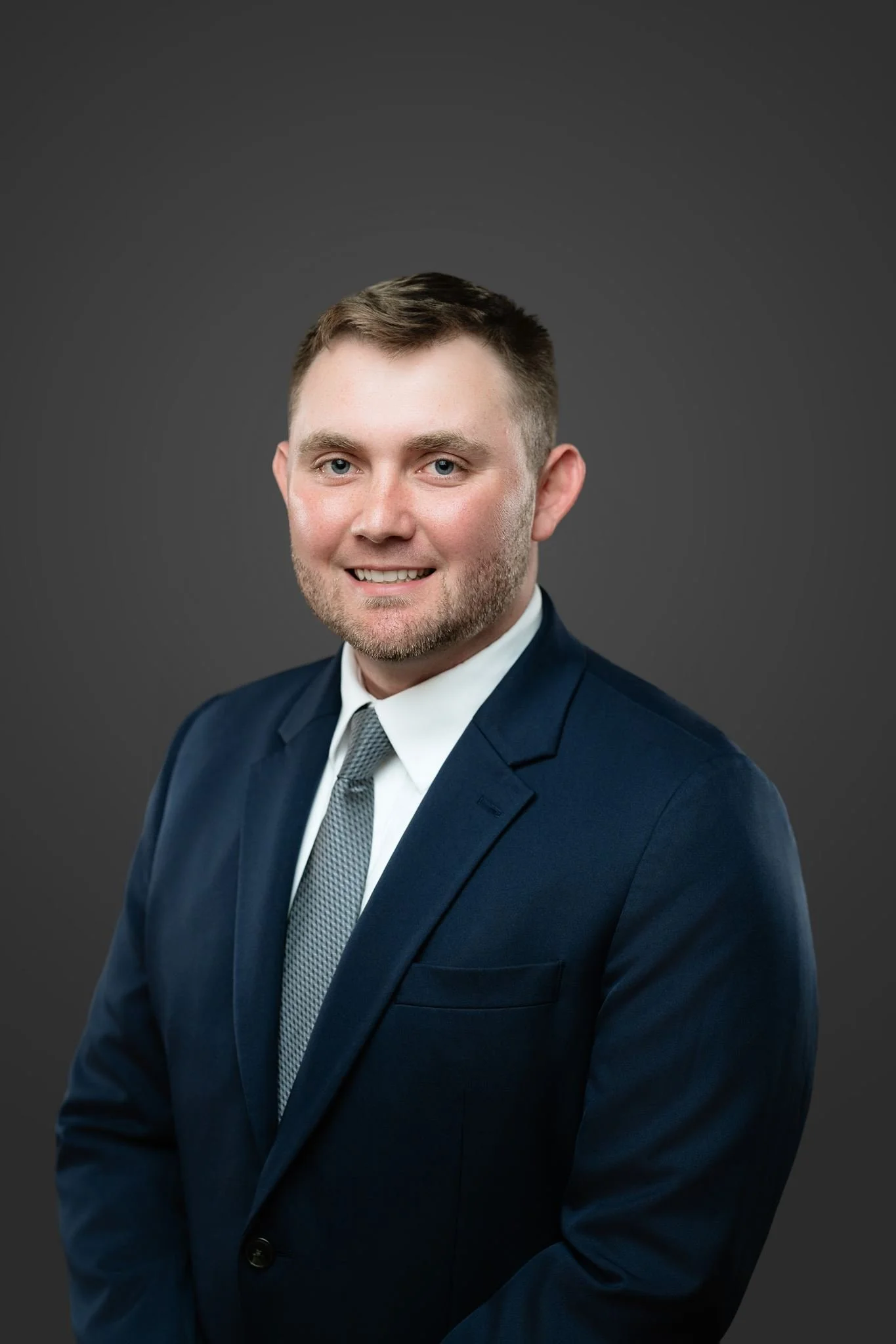 A young man with short brown hair and blue eyes, wearing a navy blue suit, white shirt, and gray tie, smiling at the camera against a dark gray background.