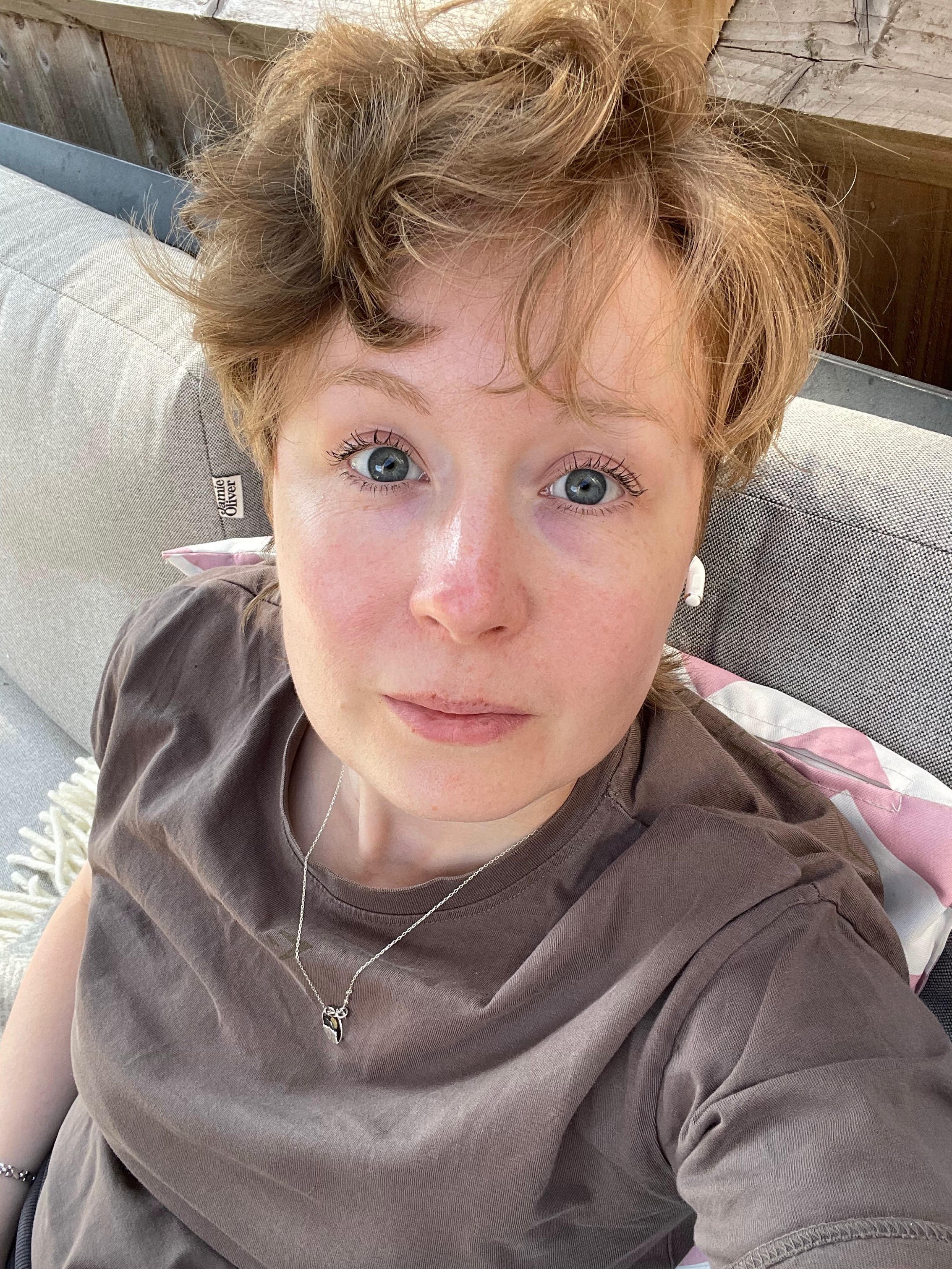 A person with short wavy red hair and blue eyes lying on a gray couch with a beige pillow behind them, wearing a brown T-shirt, a silver necklace with a pendant, and white wireless earbuds.