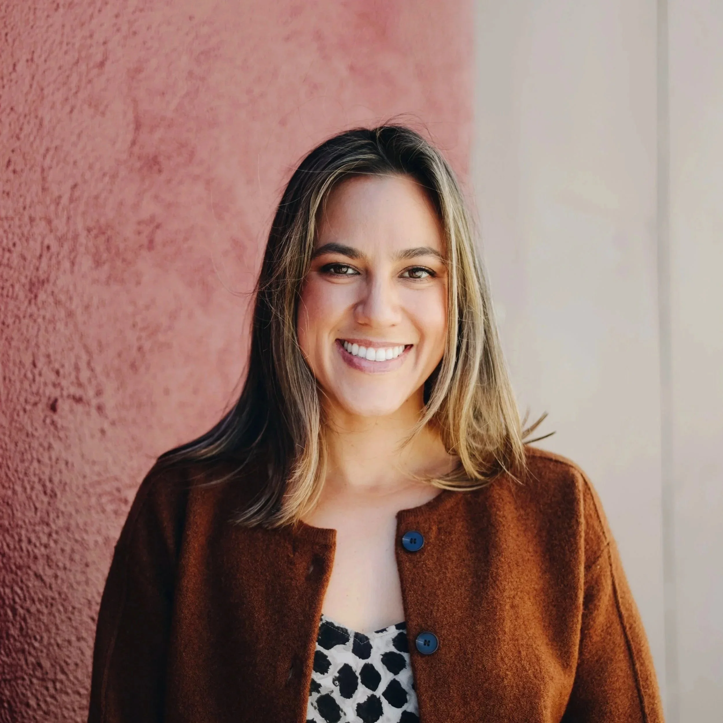 A woman named Jennifer Wong with shoulder-length blonde hair, smiling, wearing a brown jacket with blue buttons, and a white shirt with black spots. The background includes a pink wall and a white wall.