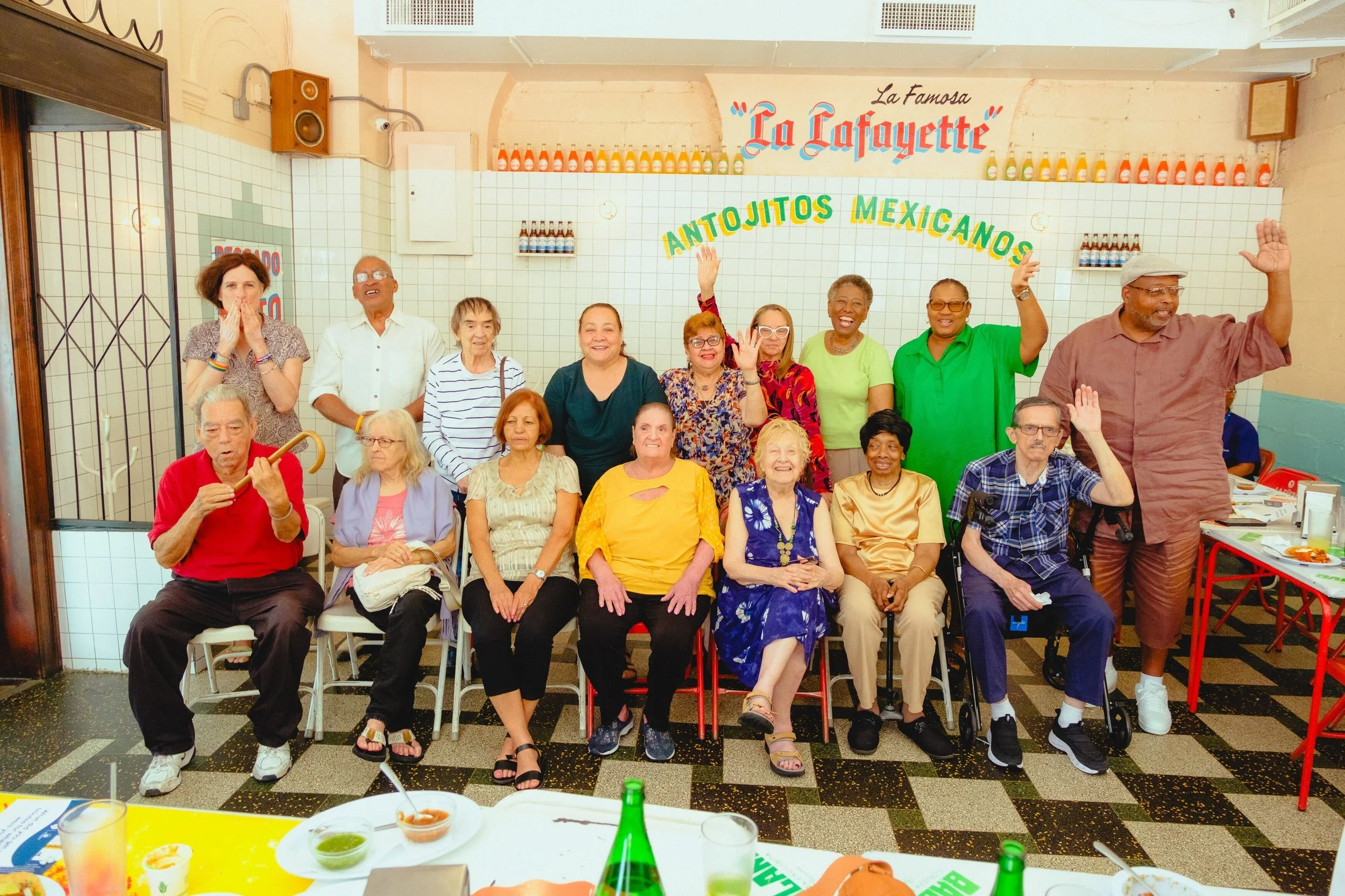 A group of diverse elderly people gathered in a restaurant, some seated and some standing, smiling and waving at the camera. The background shows a sign that says 'La Famosa La Cafayette' and 'Antojitos Mexicanos.' There are tables with food and drinks in the foreground.