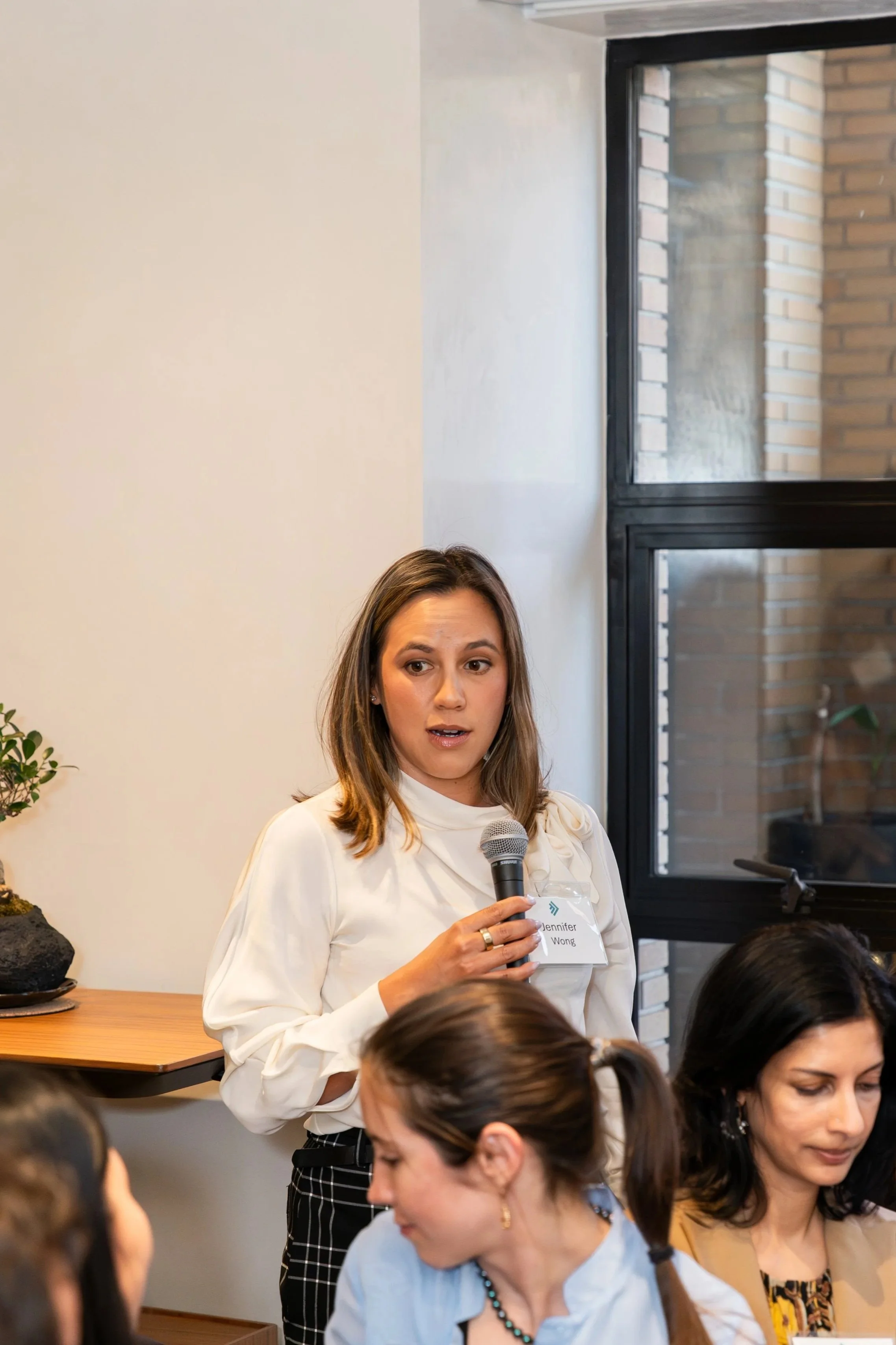 A woman named Jennifer Wong speaking into a microphone during a meeting or conference, with a group of people seated in front of her, indoors with a modern brick and window background.
