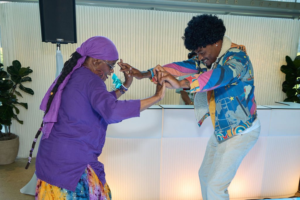 Two people dancing and smiling indoors, with a woman wearing a purple headscarf and colorful skirt, and a man with curly hair wearing a colorful patterned jacket and white pants.