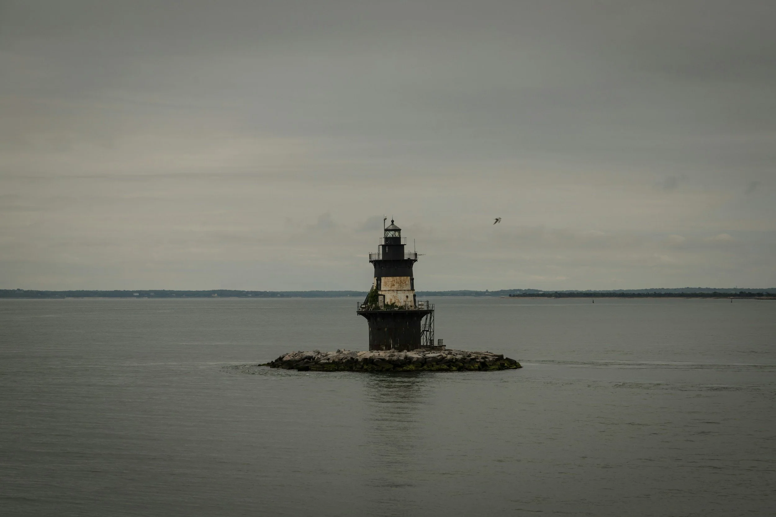 A lighthouse on a small rocky island in calm water under an overcast sky, with a bird flying nearby.