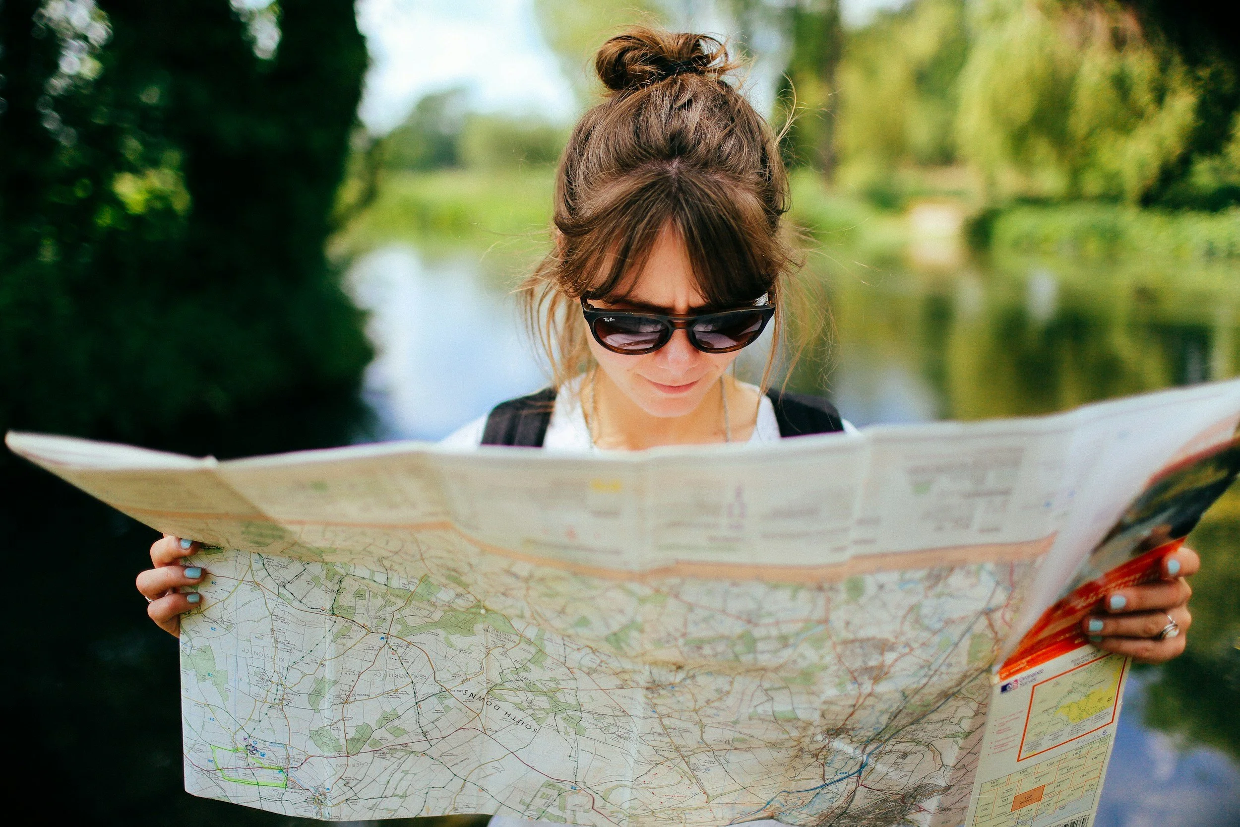 A woman with brown hair tied in a messy bun, wearing black sunglasses, is looking at a large folded map outdoors in a green, nature setting during the day.