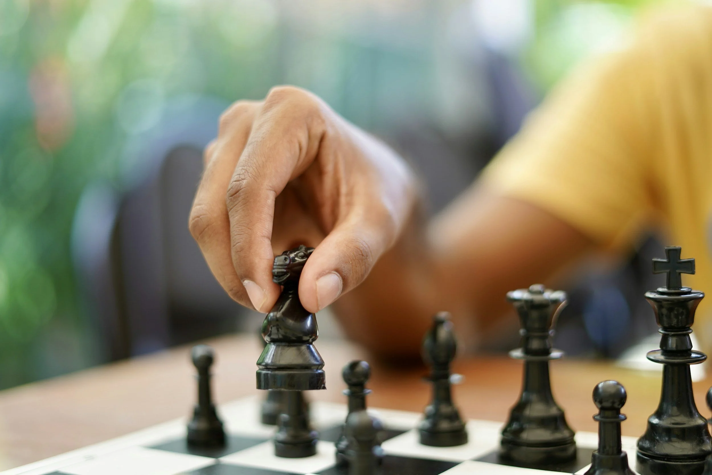 A person in a yellow shirt playing chess, moving a black piece on a chessboard against a colorful, blurred background.