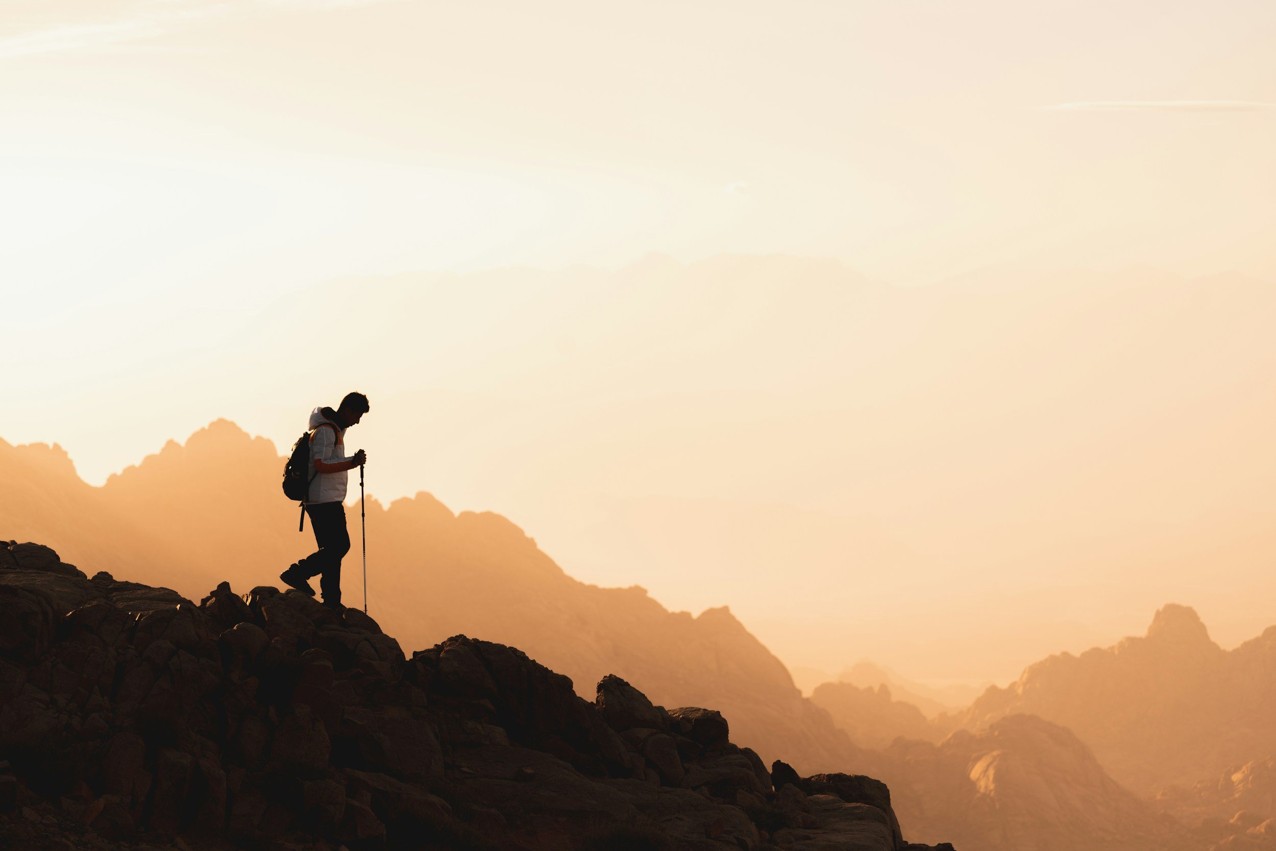 A person hiking on rocky terrain at sunrise or sunset, with mountains in the background and a glow in the sky.