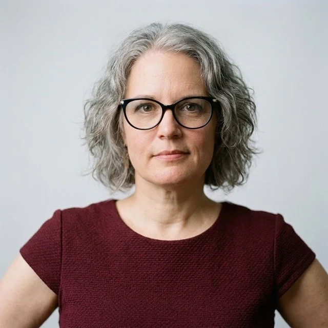 A woman with gray, curly hair and black glasses, wearing a maroon top, looking directly at the camera against a plain gray background.