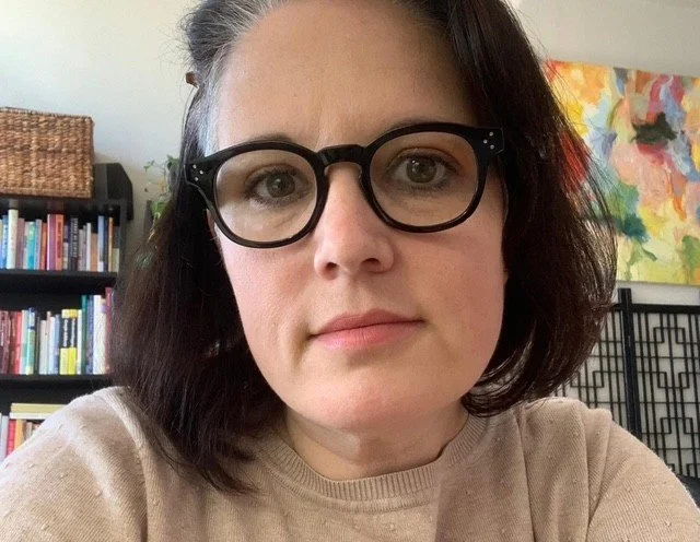 A woman with dark hair, glasses, and a beige sweater, in a room with bookshelves and colorful artwork behind her.