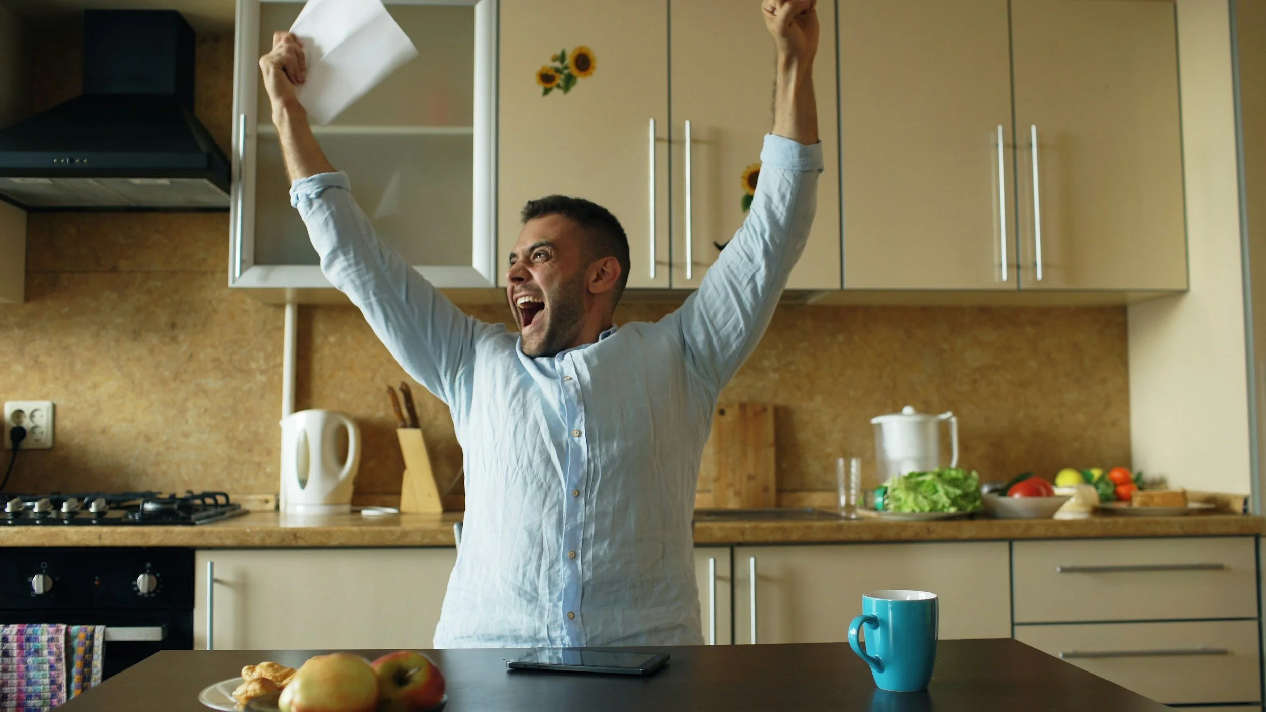A man with a joyful expression raising his arms in celebration in a kitchen, with a stove, cabinets, and various food items on the counter.
