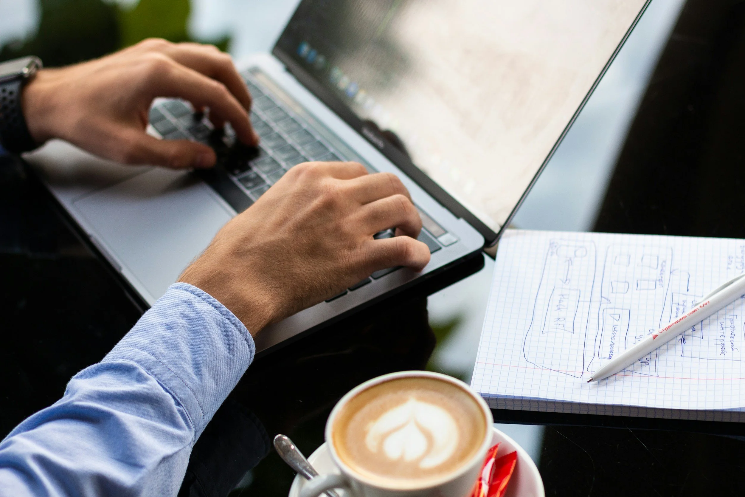Close-up of hands typing on a laptop keyboard next to a notebook with sketches, a pen, and a cup of coffee with latte art on a black table.