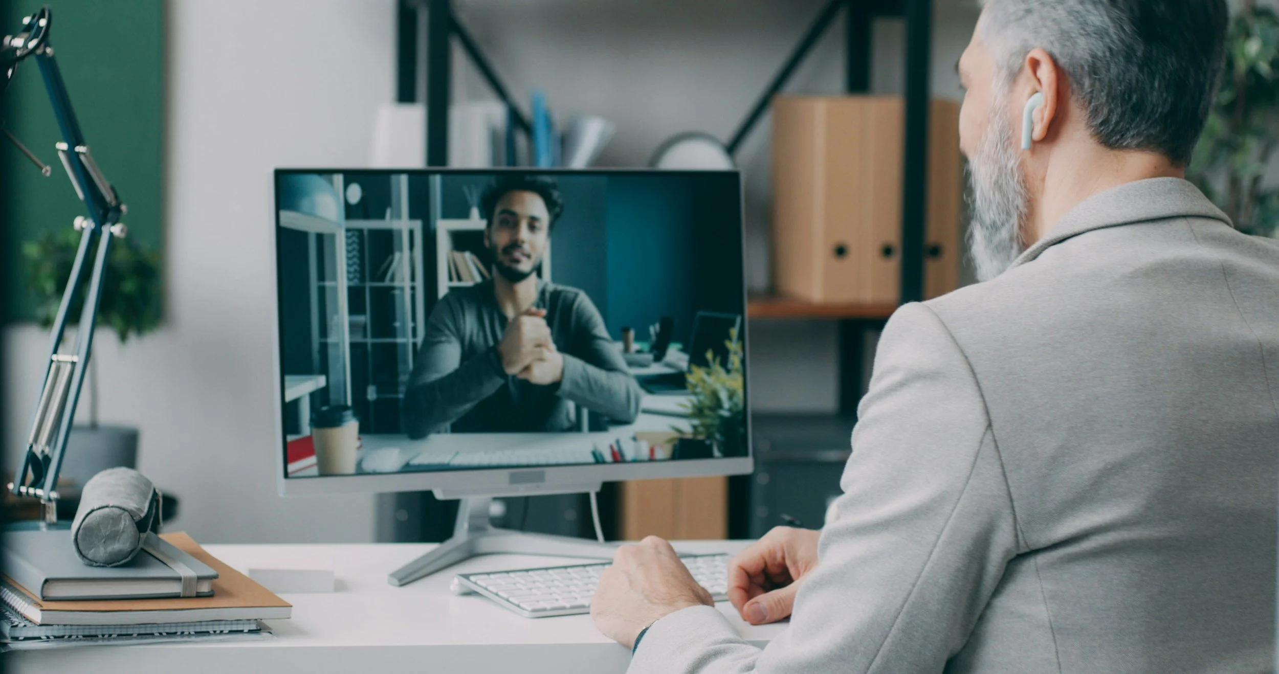 An older man with gray hair and beard, wearing gray blazer and wireless earbuds, participating in a video call on a desktop computer. The computer screen shows a young man with dark hair and beard, sitting in a well-organized room with bookshelves.