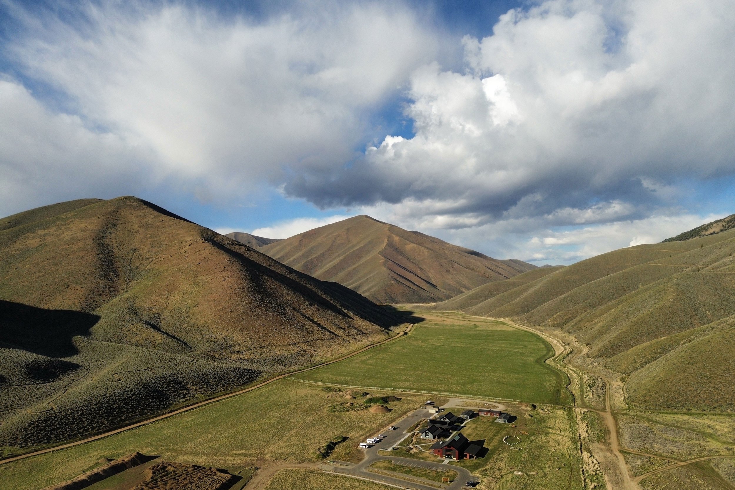 A mountainous landscape with rolling green hills under a partly cloudy sky. There are a few buildings and parked cars at the base of the hills, and winding dirt paths through the terrain.