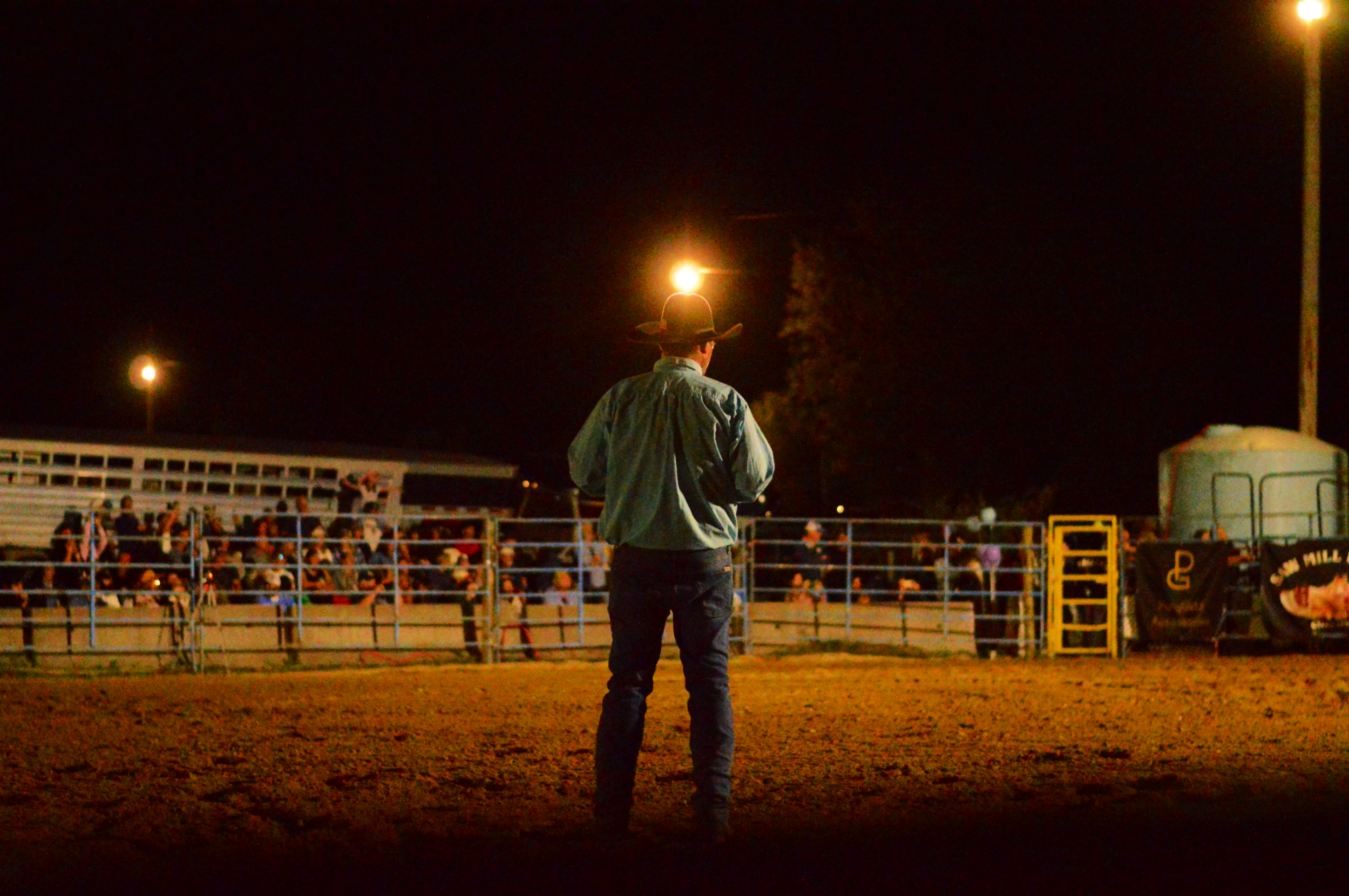 Person wearing a cowboy hat and plaid shirt standing in front of a rodeo arena at night, with a large crowd seated in the bleachers in the background.