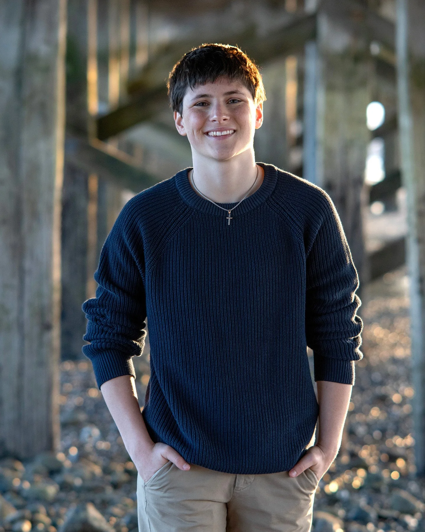 A young person with short dark hair smiling, standing outdoors under a bridge during sunset, wearing a navy blue ribbed sweater, tan pants, and a cross necklace.