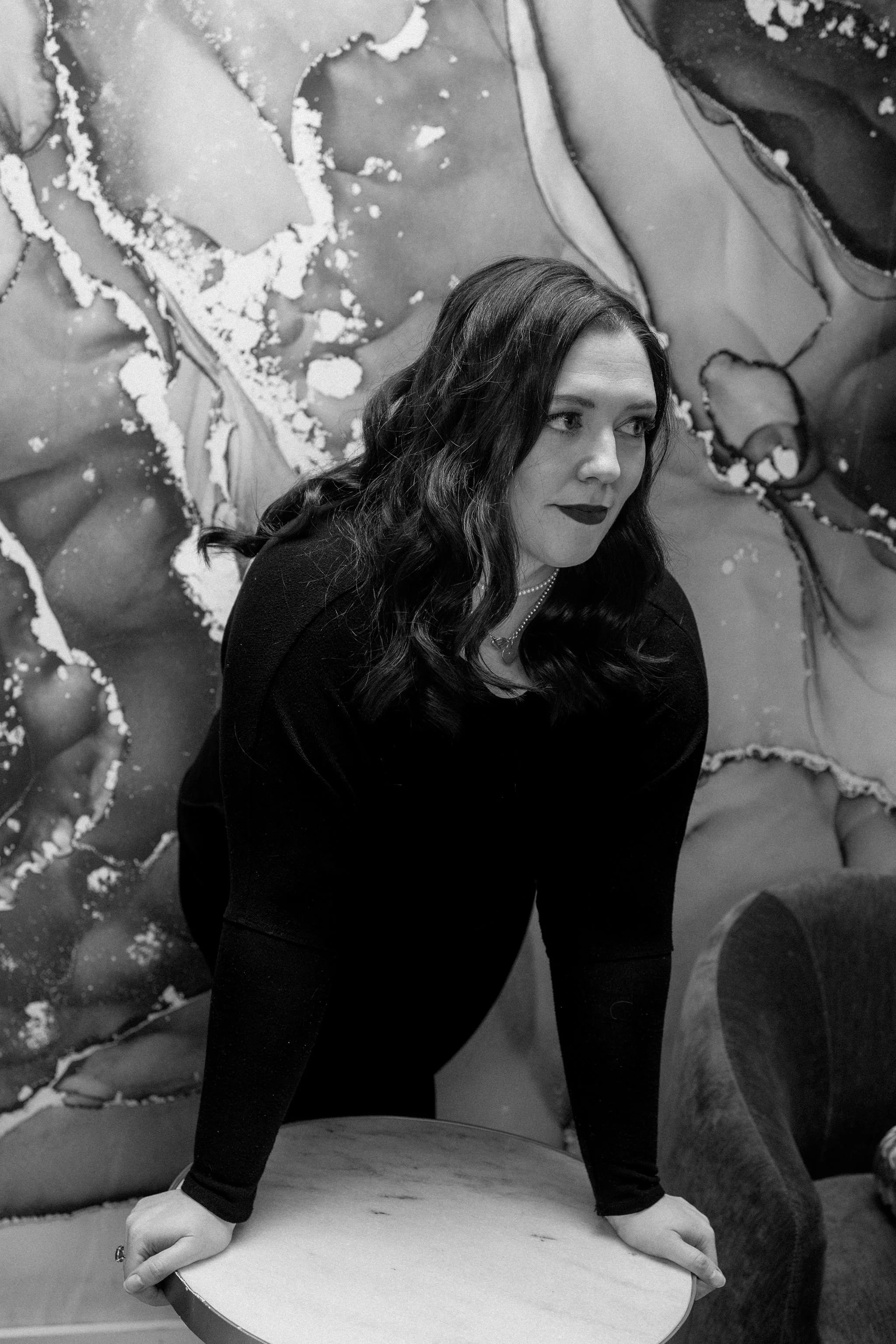 A woman with long dark hair, dressed in black, stands in front of a colorful abstract mural. She has makeup with dark lipstick and appears to be looking off to the side. She is resting both hands on a rounded table or surface.
