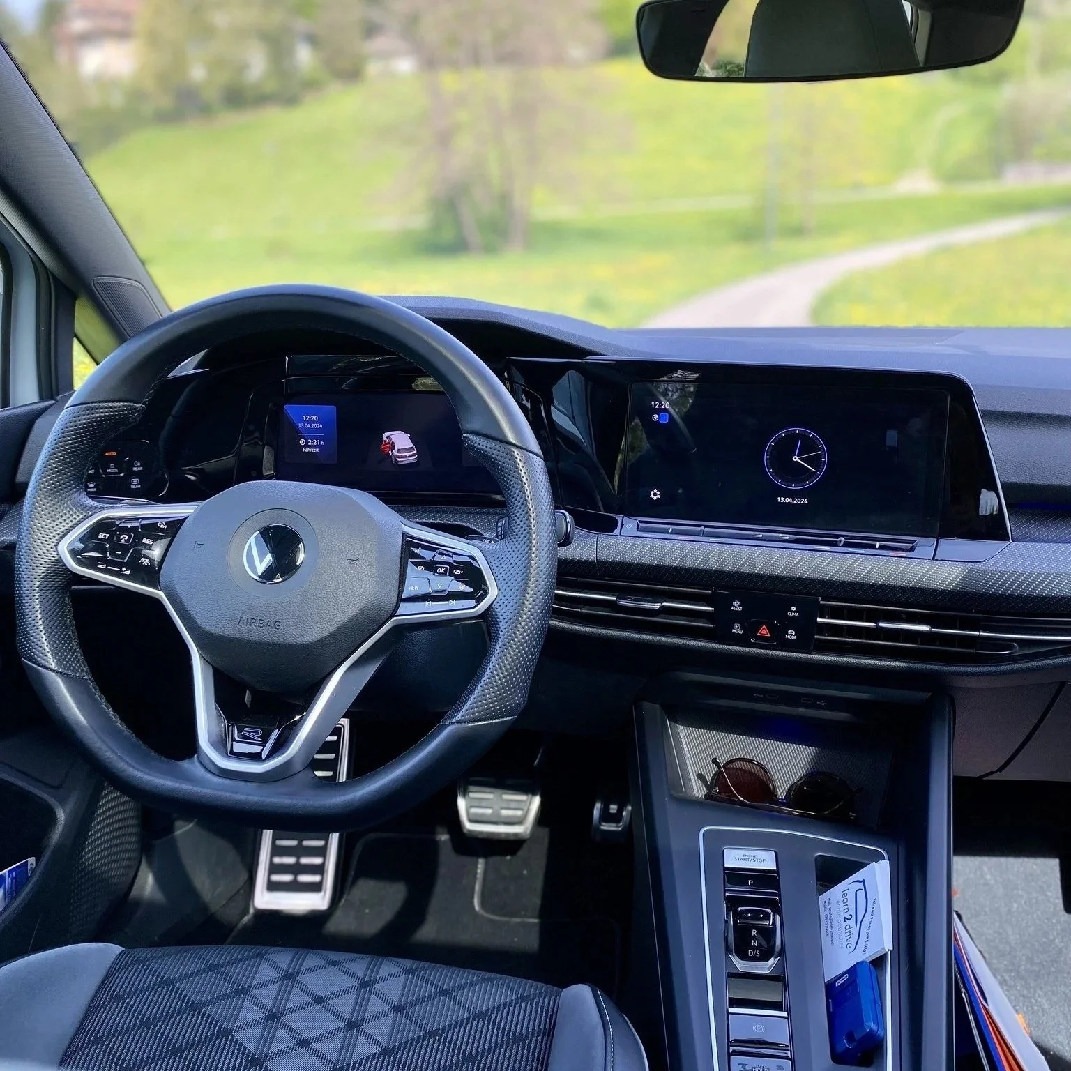 Innensicht eines modernen Autos mit digitalem Armaturenbrett, einem schwarzen Lenkrad, einem großen Display über der Mittelkonsole, in einem grünen Landschaftsgebiet.