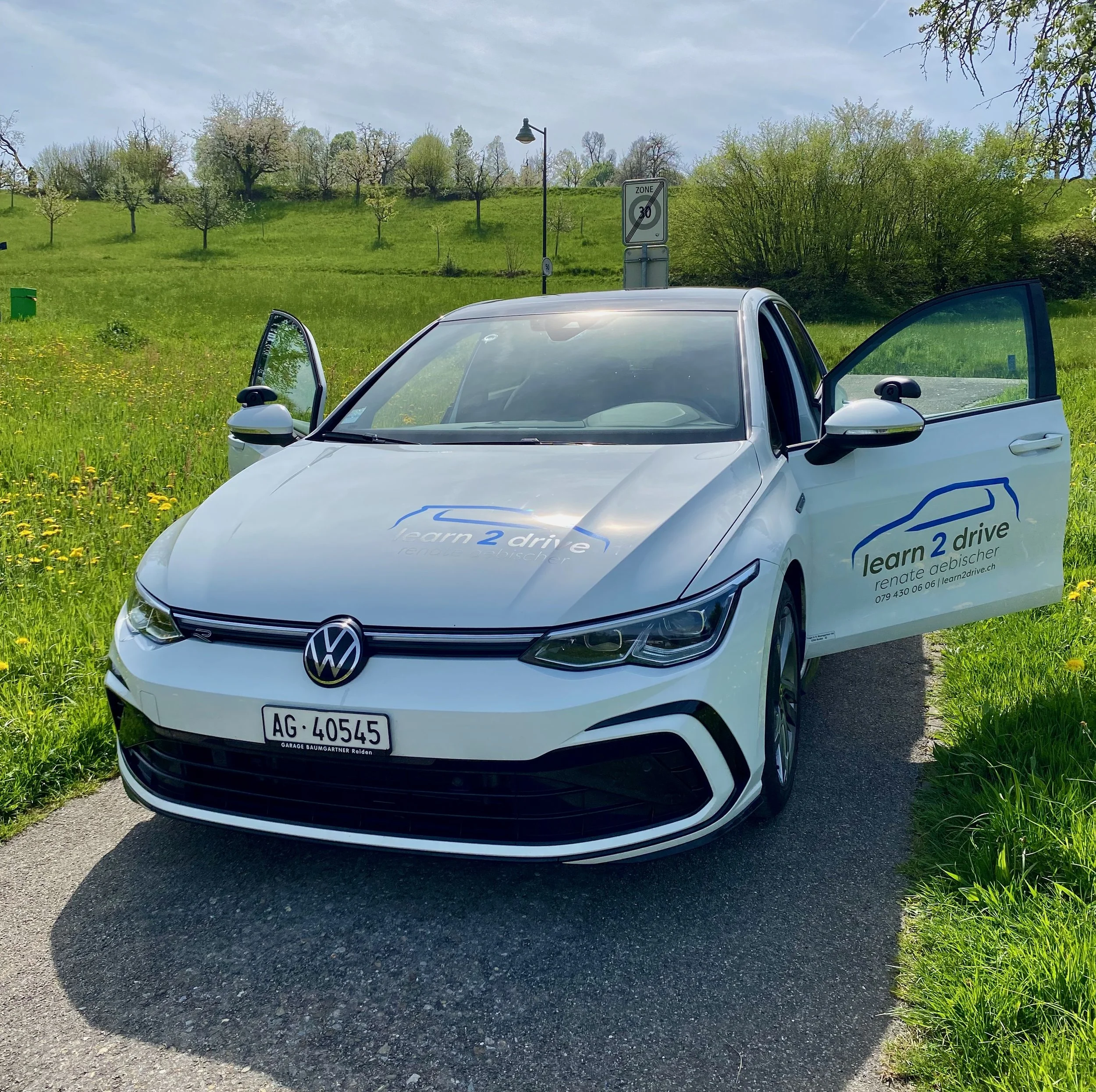Ein weißes Auto, vermutlich ein Elektrofahrzeug, steht auf einem Landweg. Es hat Aufkleber mit der Aufschrift "learn 2 drive" und ist an einer grünen Wiesenfläche und Bäumen zu sehen. Im Hintergrund gibt es eine Hügellandschaft mit Bäumen und eine Straße mit Verkehrszeichen.