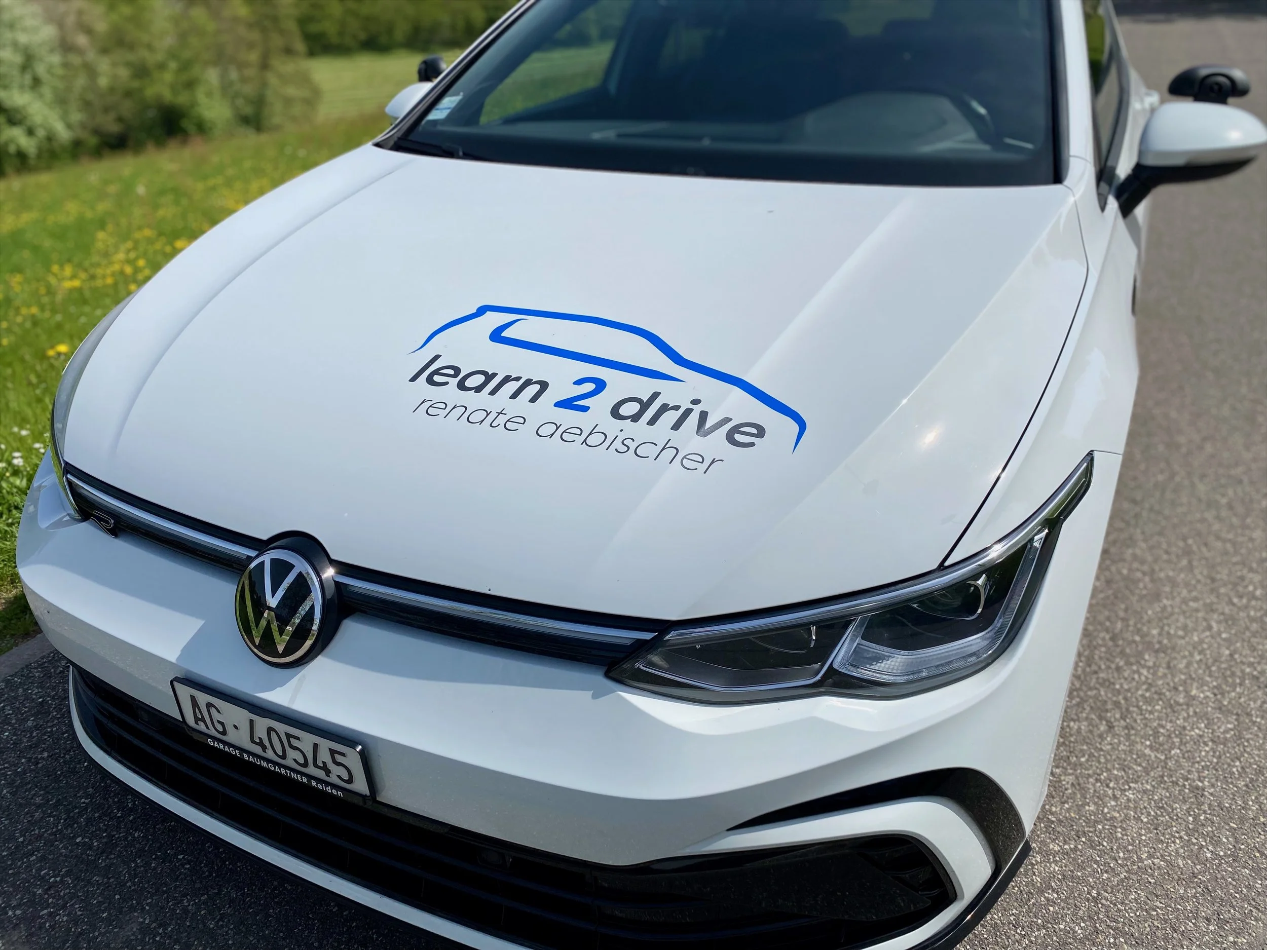 Ein weißen Volkswagen-Auto mit dem Logo 'learn2drive' auf der Motorhaube, das auf einer Straße parkt, mit grünen Bäumen im Hintergrund.