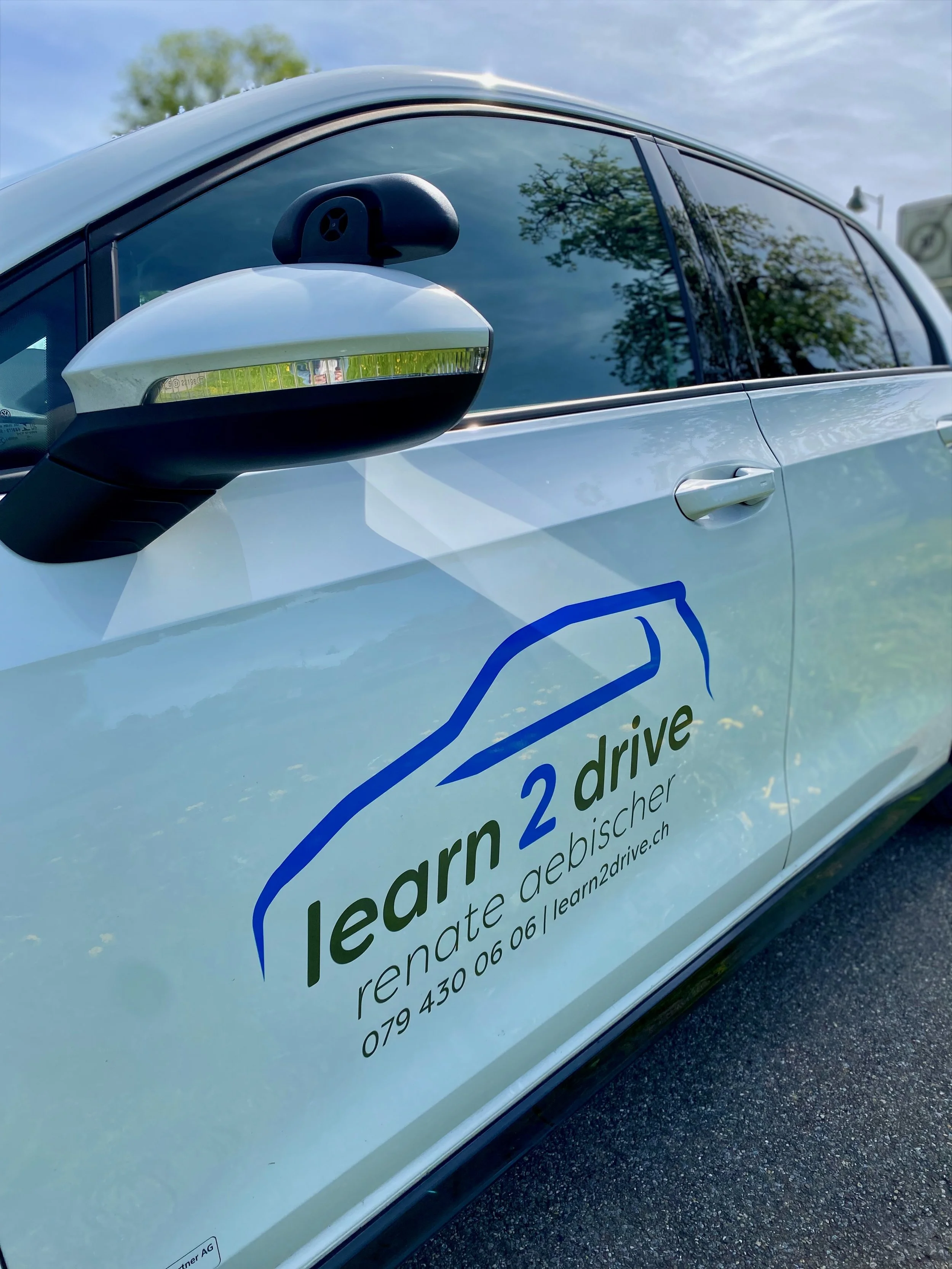 Ein weißes Auto mit einem Logo und Text für eine Lernfahrseite. Der Text lautet: "learn 2 drive gebüscher" mit Kontaktinformationen. Im Hintergrund sind Bäume und ein blauer Himmel zu sehen.