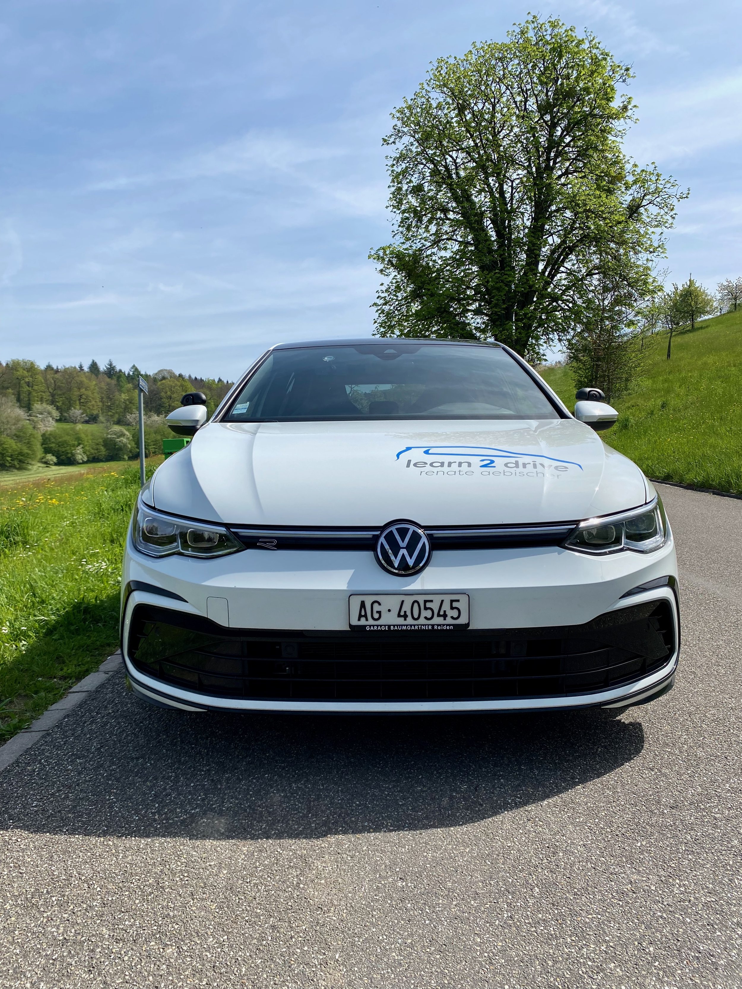Ein weißer Volkswagen E-Auto mit einem Logo 'learn 2 drive' auf der Motorhaube parkt auf einer Straße, im Hintergrund grüne Bäume und eine Wiese unter einem blauen Himmel.