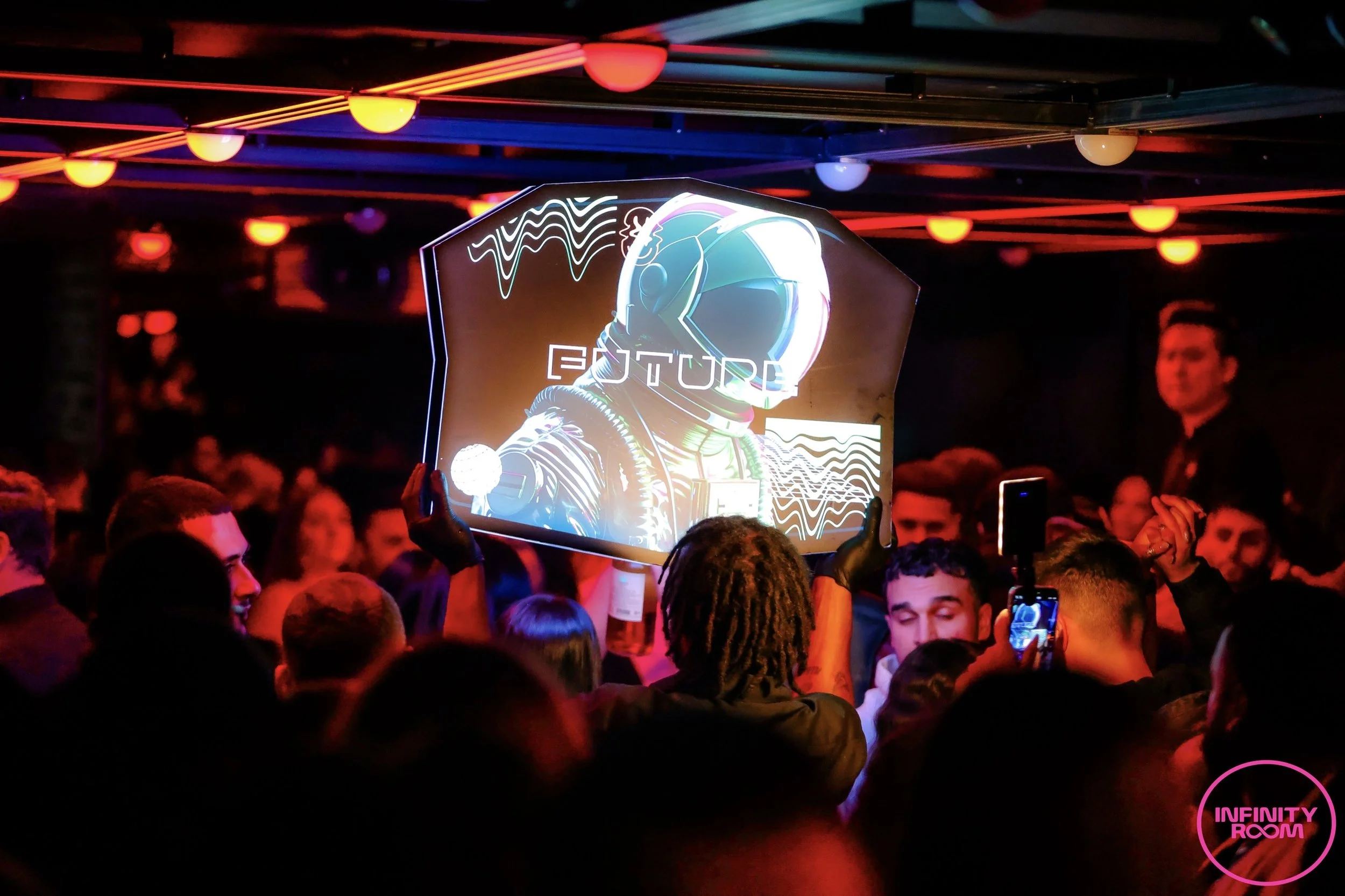 Crowd at a party or event with neon lights, a large digital display showing a futuristic astronaut helmet, and a pink neon sign reading 'INFINITY ROOM'.