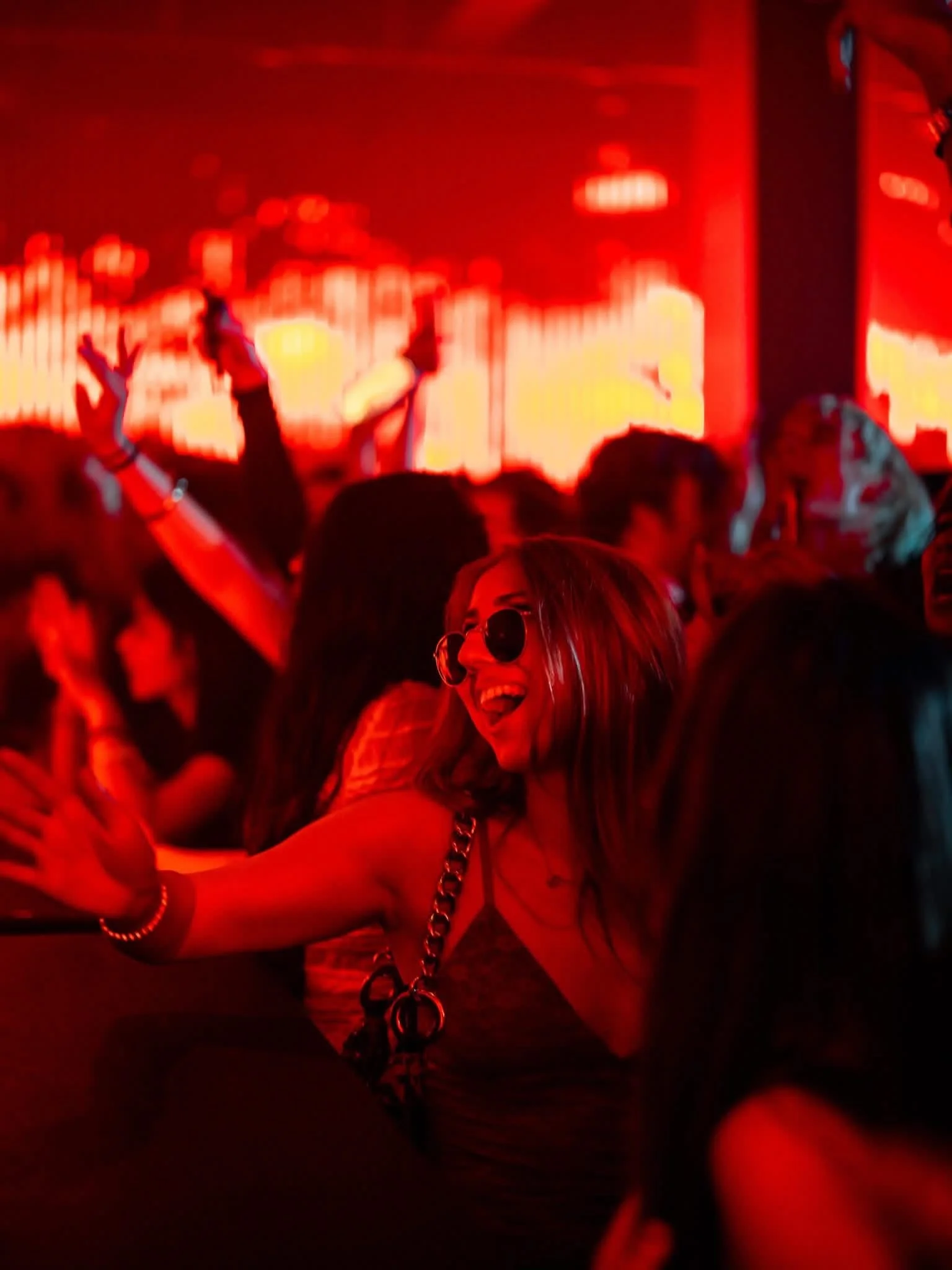 A woman with blonde hair, sunglasses, and a big smile dancing at a party with red lighting, surrounded by other people.