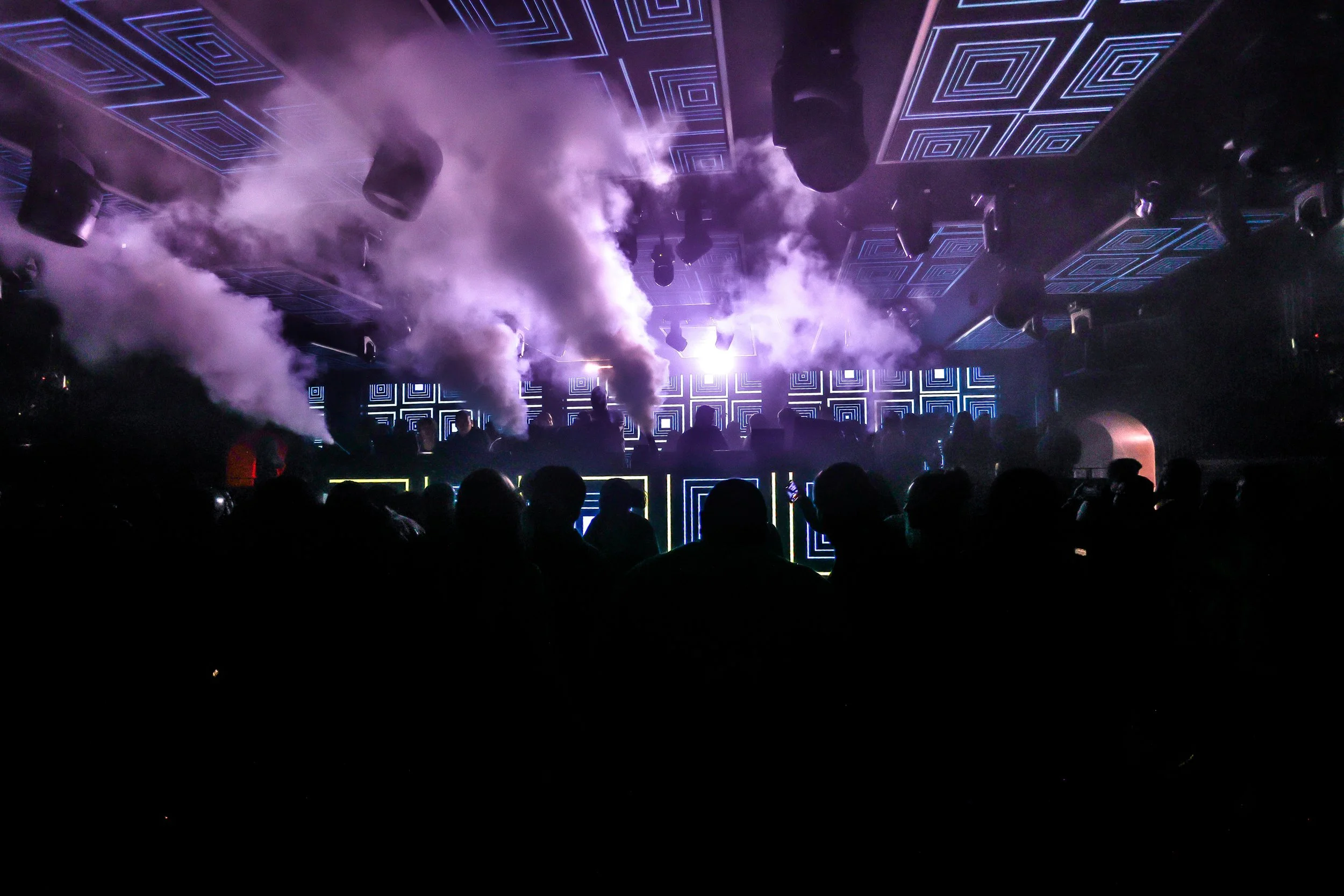 A dark nightclub with geometric light patterns on the ceiling and fog or smoke effects, with silhouettes of people dancing and socializing, and fog machines emitting clouds of smoke.