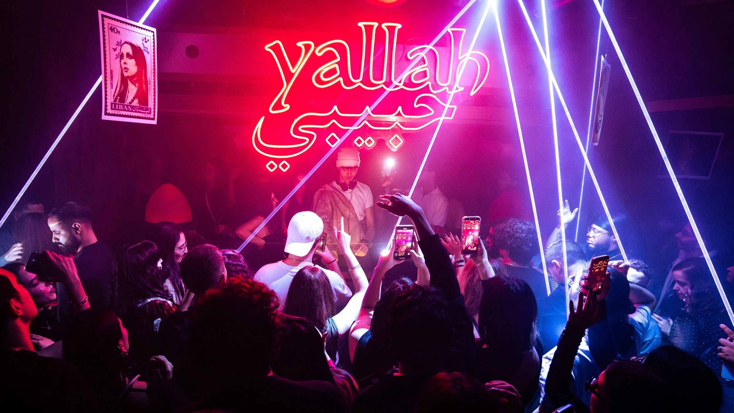A nightclub scene with a crowd dancing and taking photos, neon sign saying 'Yallah' in Arabic and English, blue and purple laser lights, and a poster of a woman on the wall.