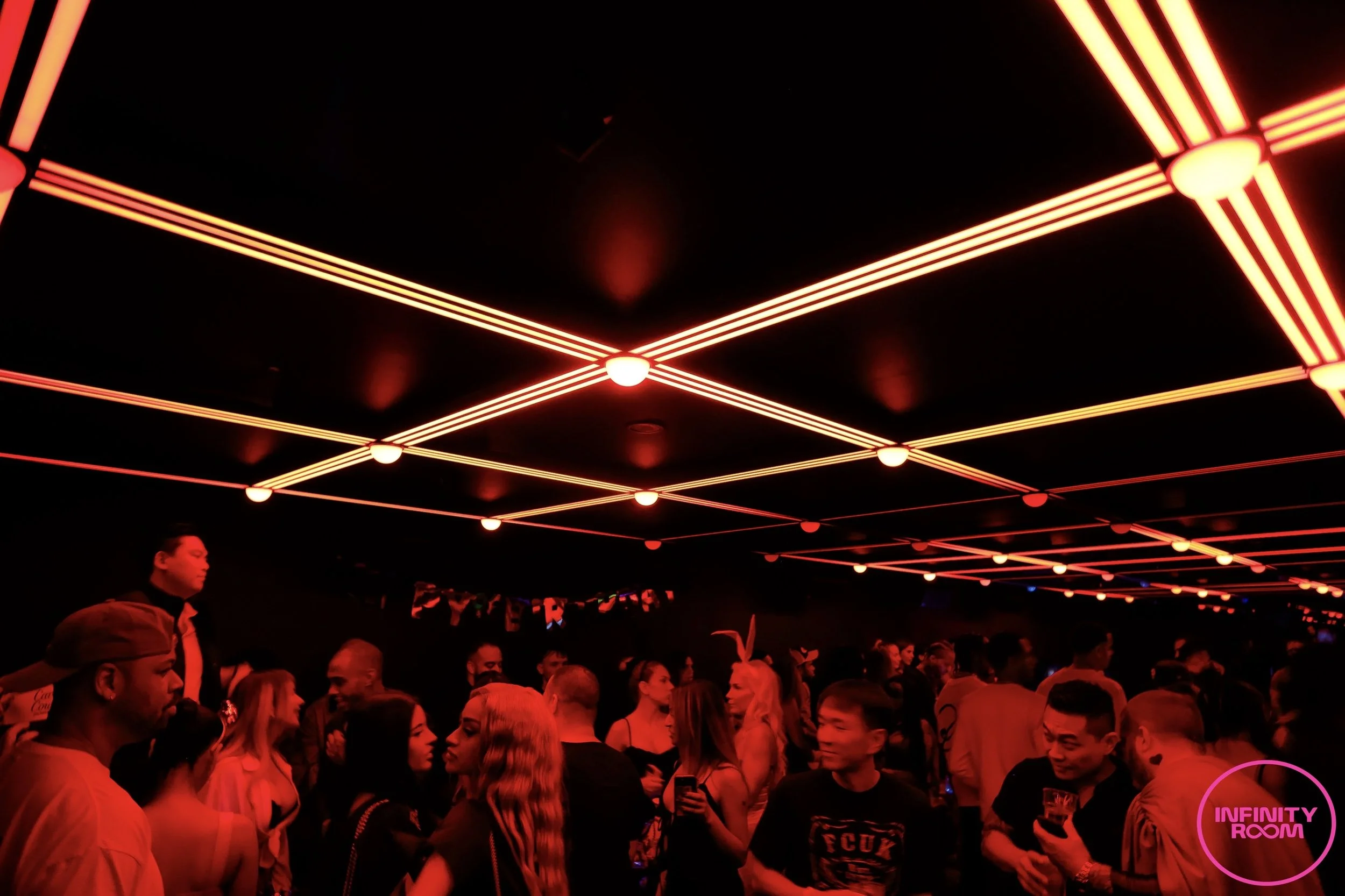 A crowded party scene with people under a ceiling decorated with red neon lights arranged in geometric patterns. There is a pink sign that reads 'Infinity Room' in the bottom right corner.