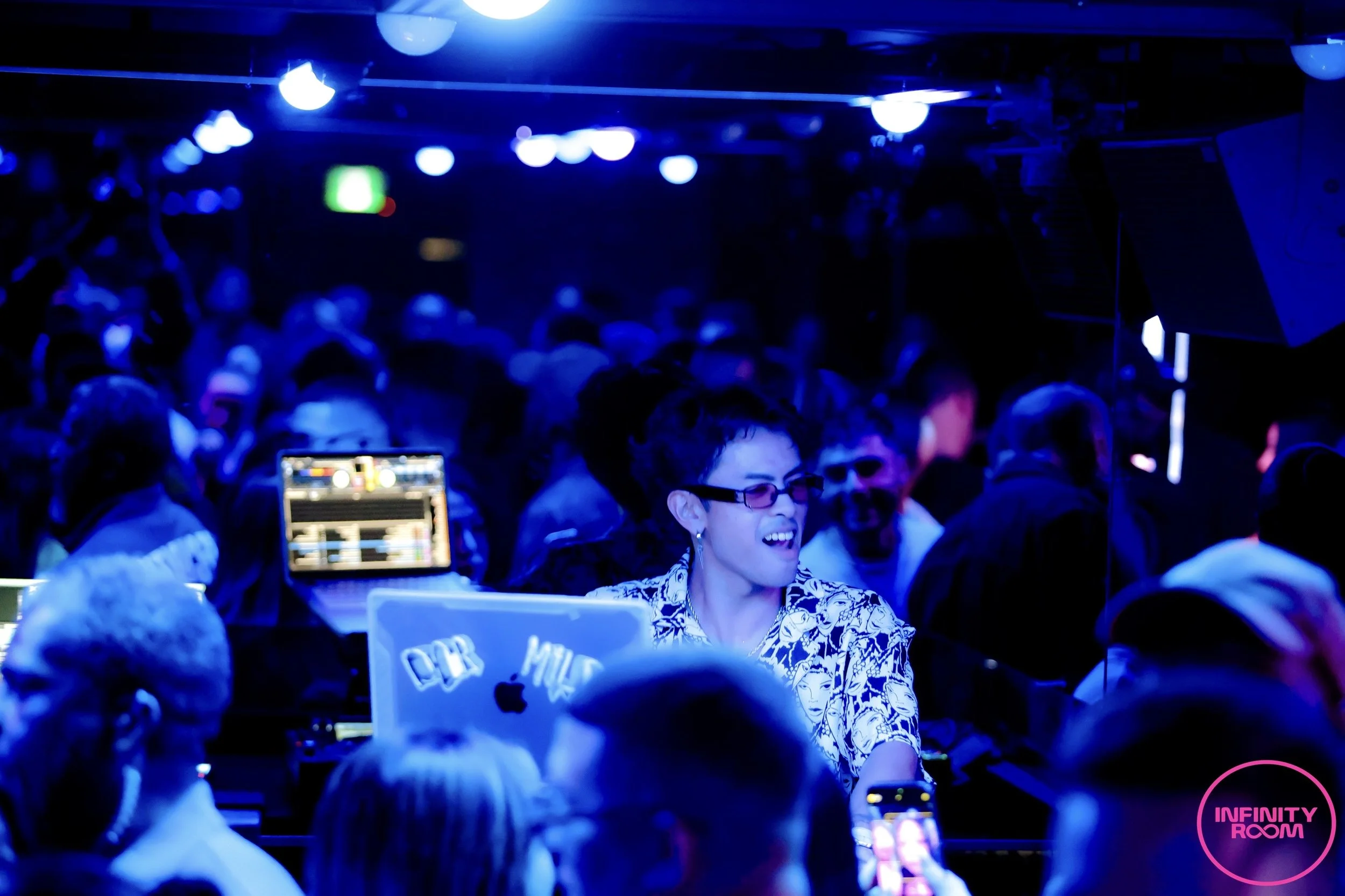 A woman with glasses and a patterned top is standing near a DJ booth with a laptop, surrounded by a crowd inside a dark, blue-lit club. The logo 'Infinity Room' is visible in the bottom right corner.