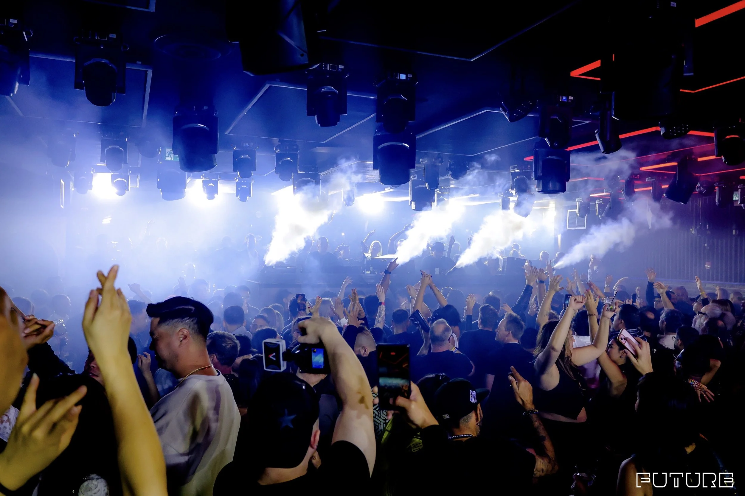 A crowded concert or nightclub scene with people dancing and taking photos, on a dark stage with bright blue and white lighting, smoke, and hanging speakers.