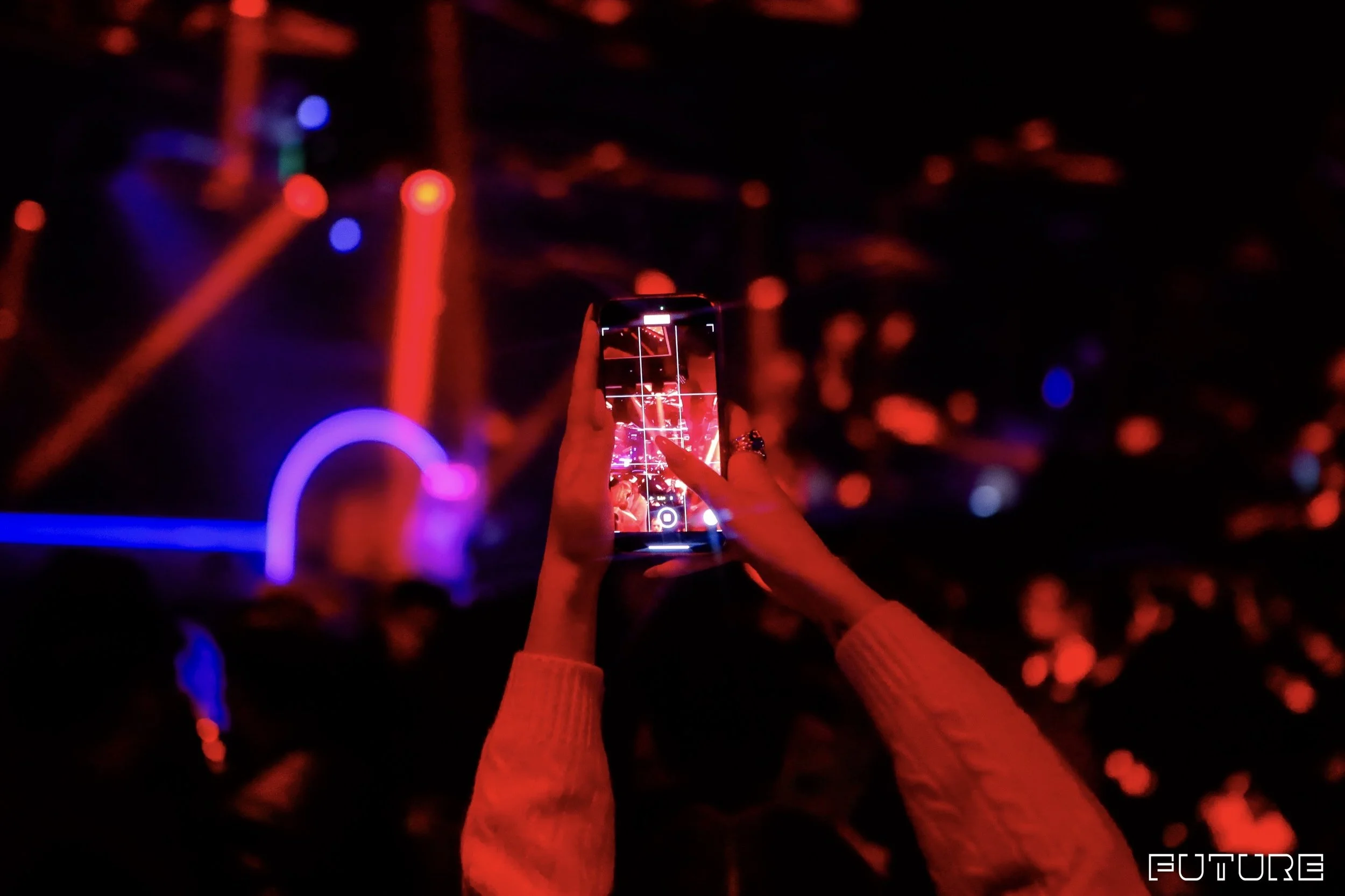 A person in a red long-sleeve shirt holds a smartphone with a grid pattern on the screen, capturing a photo of colorful, blurry neon lights in a dark setting. The lights include red, blue, and green hues, creating a vibrant background. The word 'FUTURE' is visible in white text in the bottom right corner.