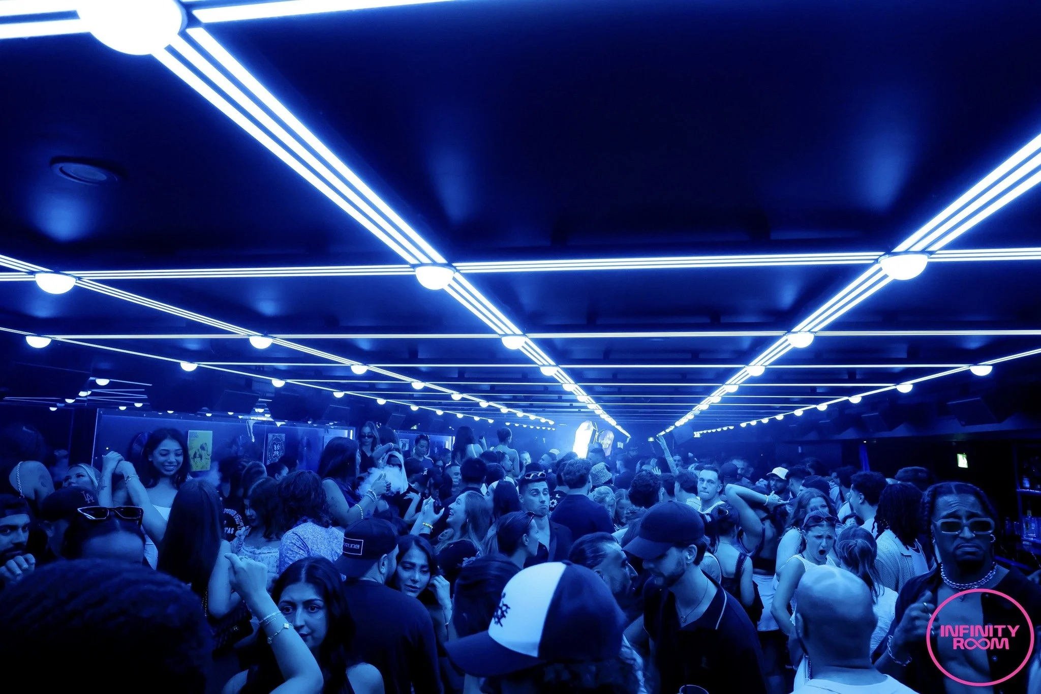 Crowd of people at an indoor event with blue LED ceiling lights. There is a pink sign reading 'INFINITY ROOM' in the lower right corner.