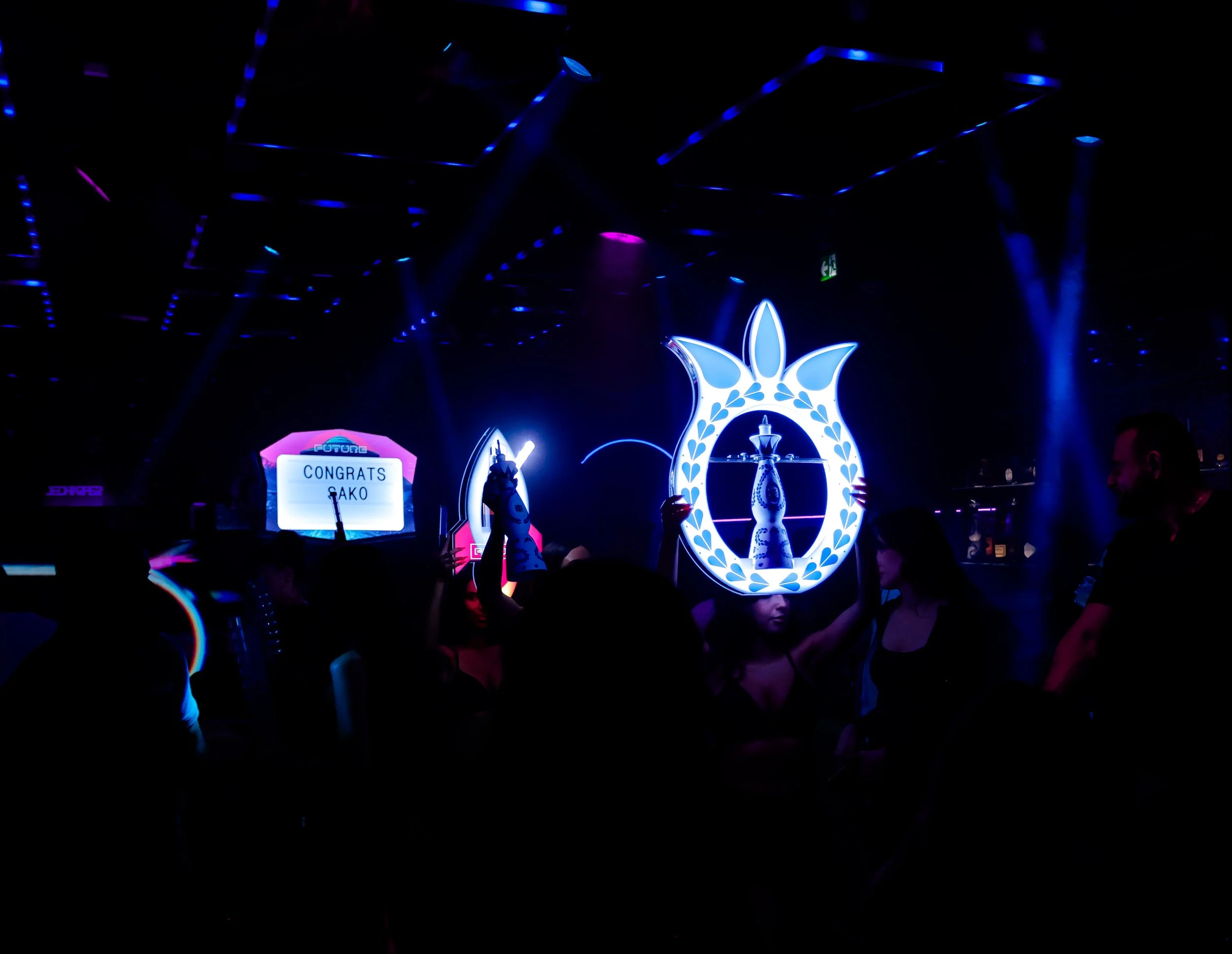 Dark nightclub scene with neon blue and pink lights, a large decorative arch with a fountain in the center, and a group of people dancing and taking pictures.