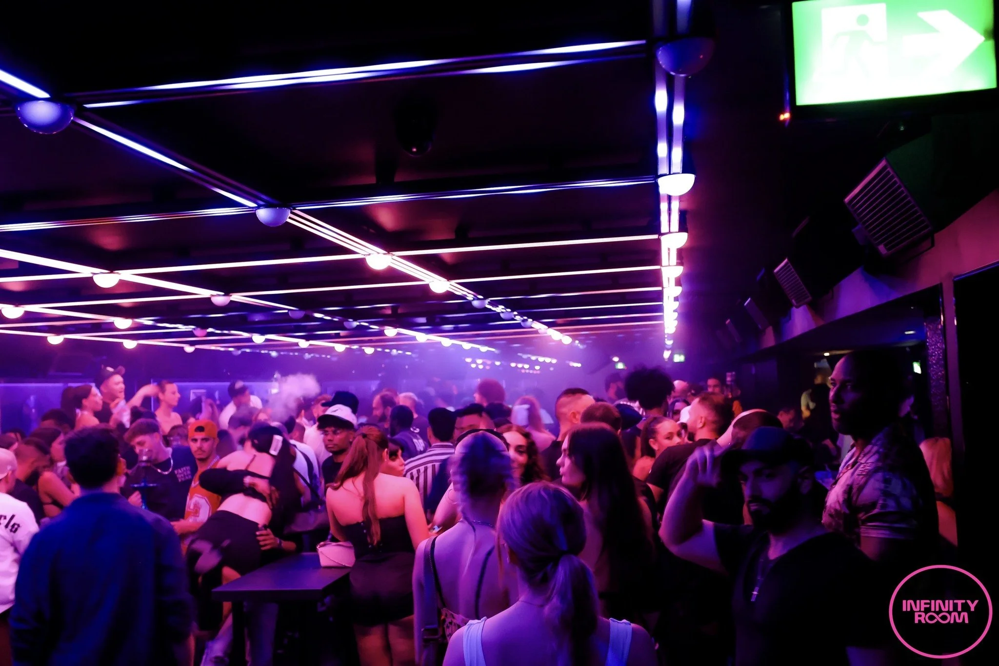 Crowd of people in a nightclub with pink and purple neon lighting and a green exit sign.