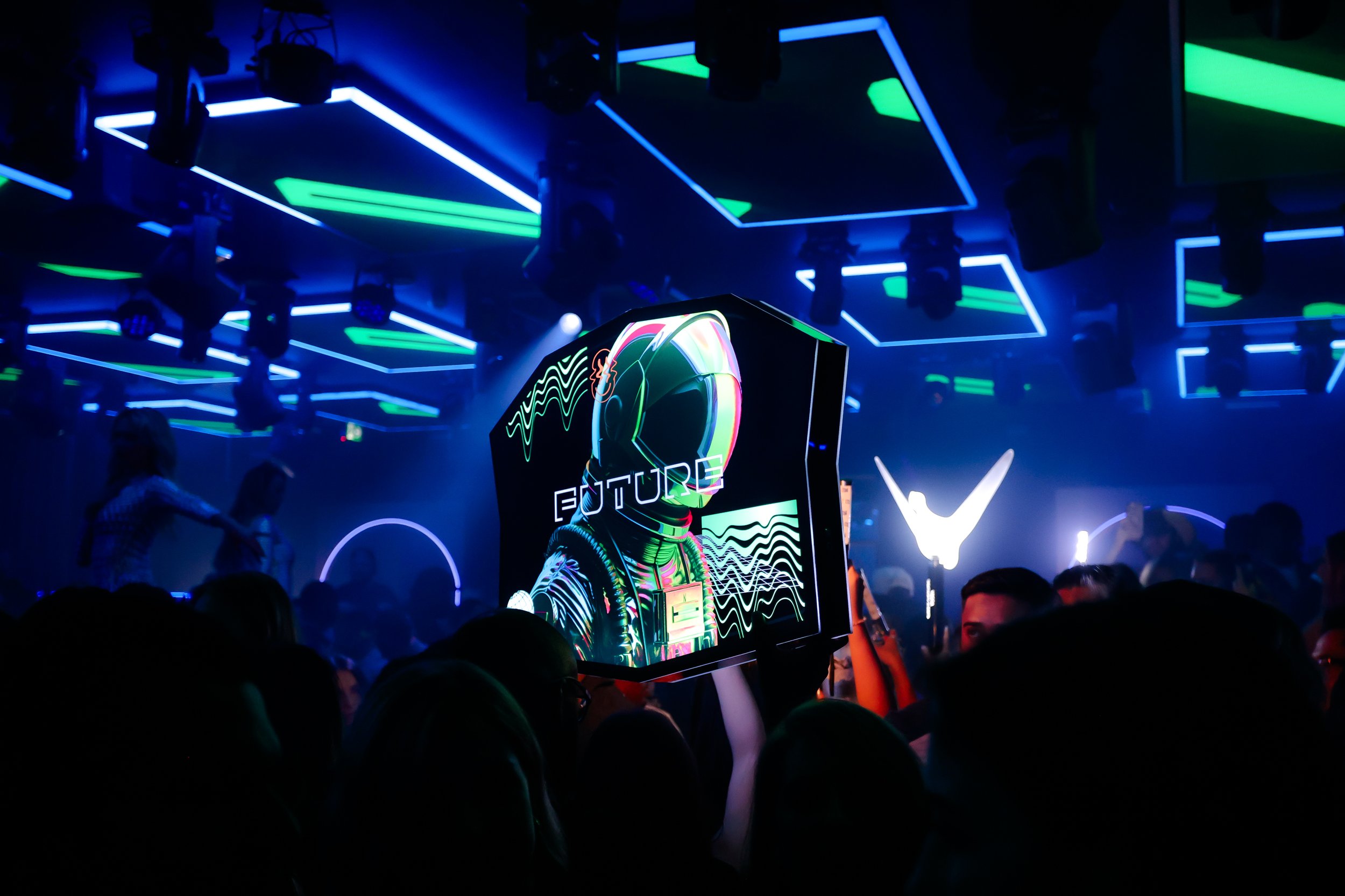 A crowded nightclub with neon blue and green lighting. A person is holding a tablet displaying digital artwork of a futuristic astronaut with the word "FUTURE" on the screen. The ceiling has geometric neon shapes, creating a vibrant atmosphere.