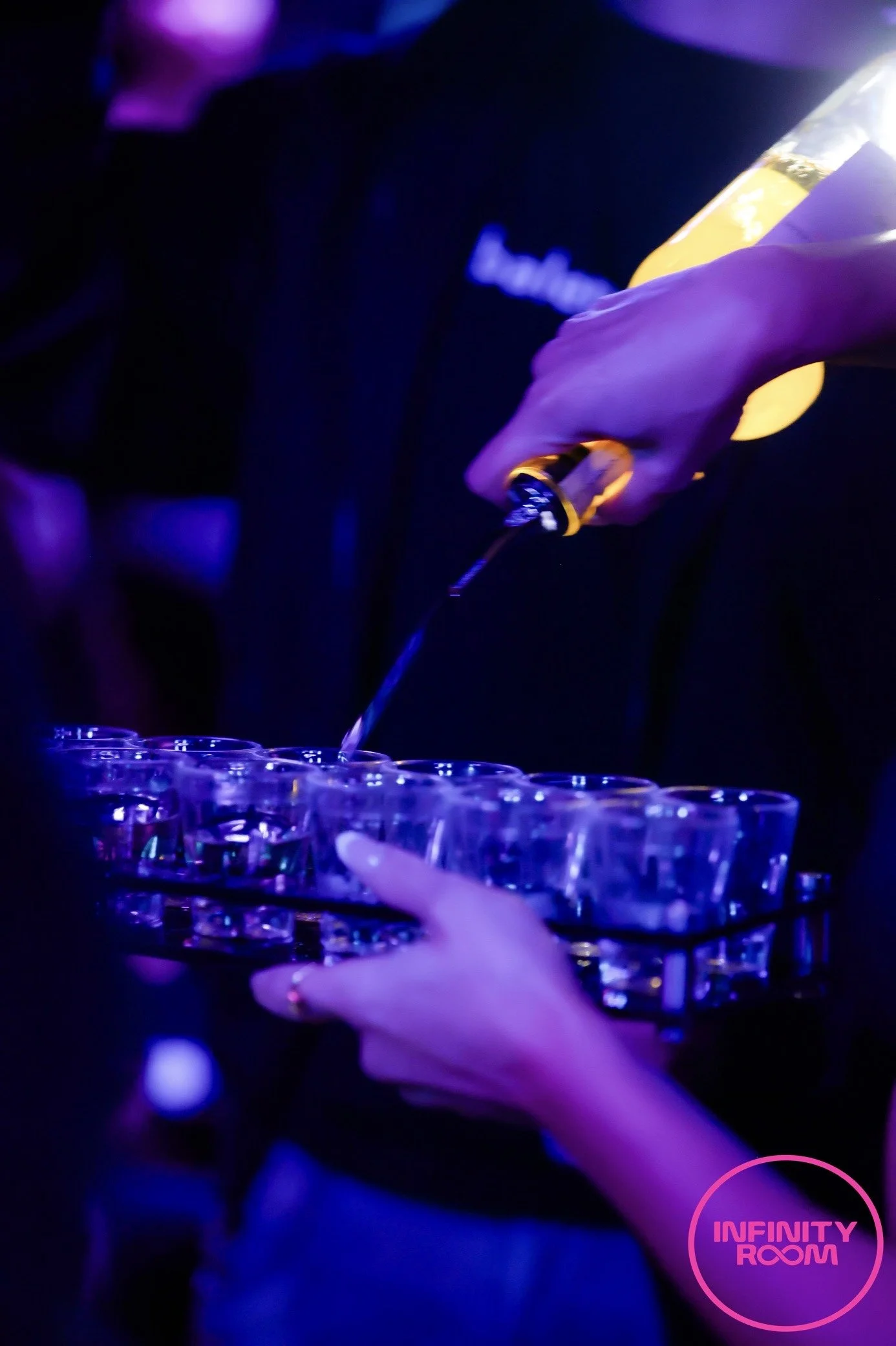 Person pouring a drink into a row of glasses at a nightclub called 'Infinity Room' with neon pink lighting.