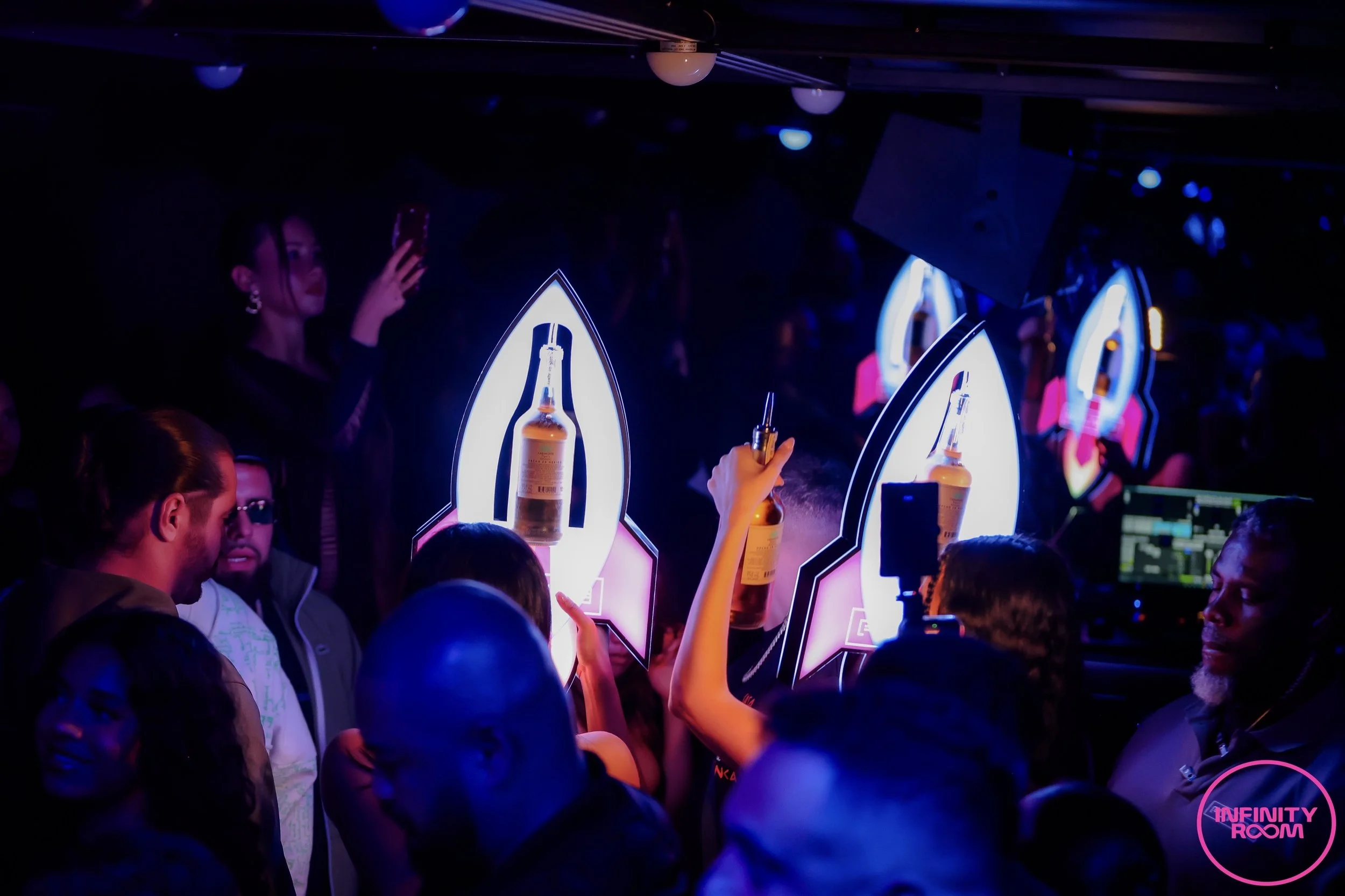 A crowded nightclub scene with people celebrating and holding bottles of liquor towards illuminated signs, including one labeled 'Infinity Room' in pink. The scene features vibrant lighting with neon accents, creating an energetic atmosphere.