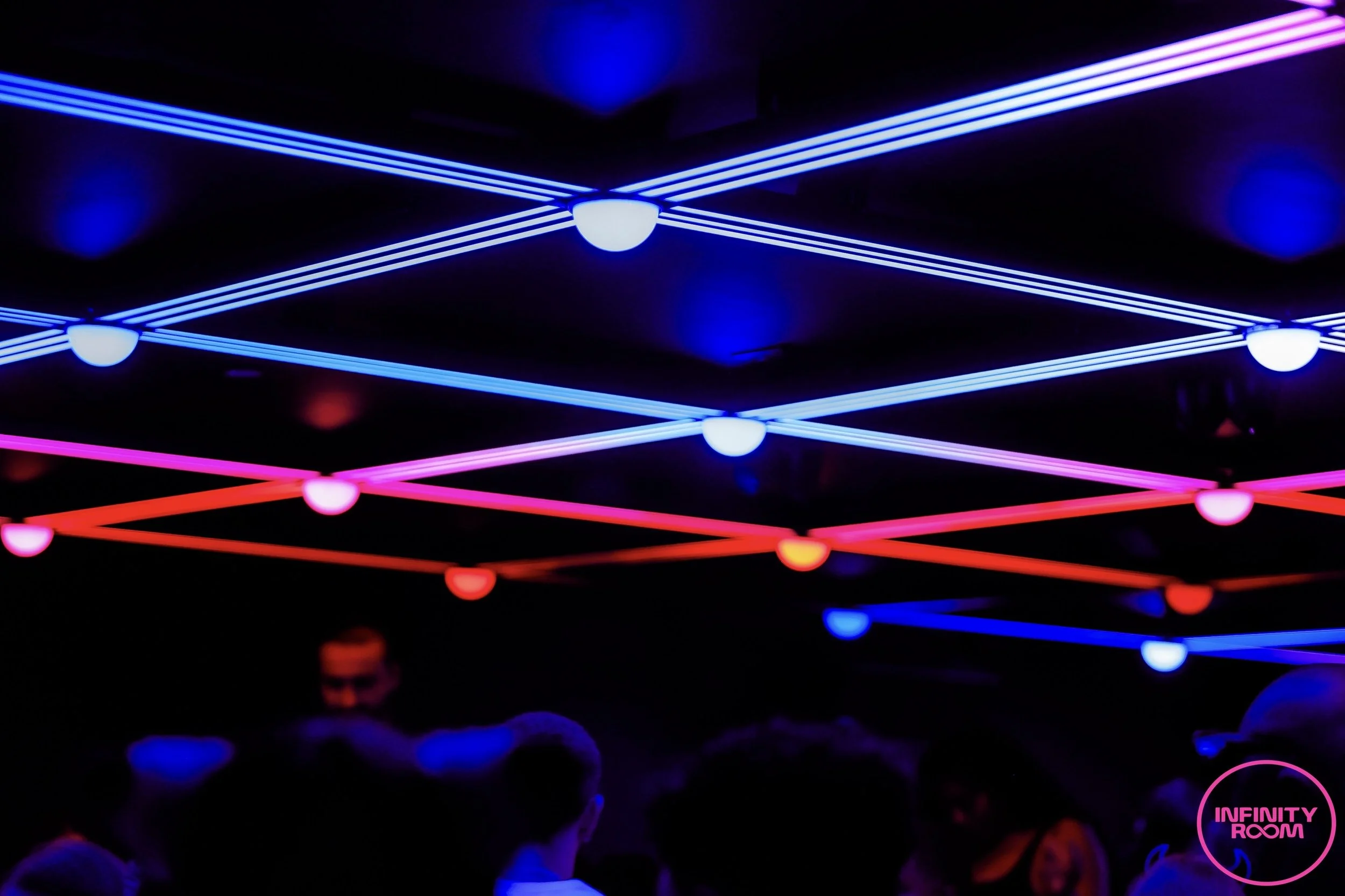 Colorful neon light installation with intersecting blue, pink, and red lines in a dark room, with a crowd of people visible at the bottom and a pink circle with text 'INFINITY ROOM' in the lower right corner.