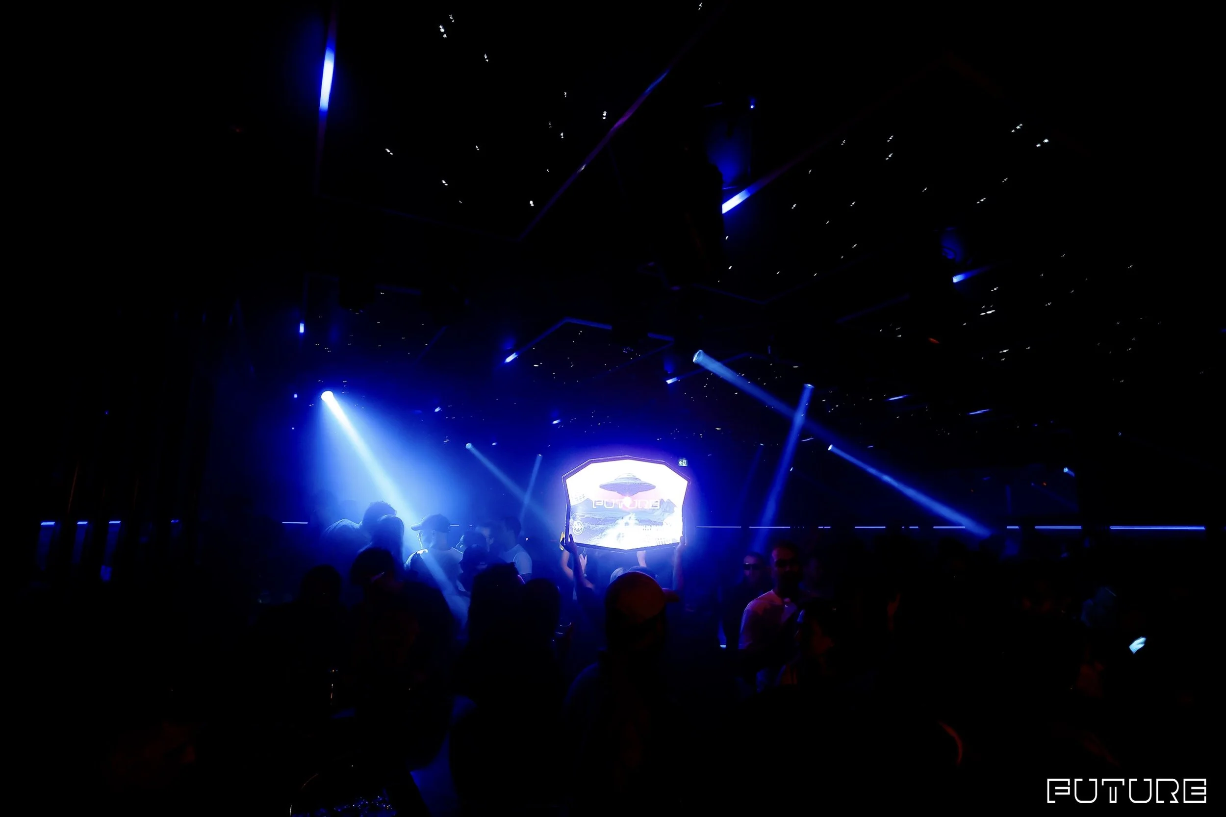 A dark nightclub with blue and purple stage lighting, silhouettes of people, and a large screen displaying the word "Future".