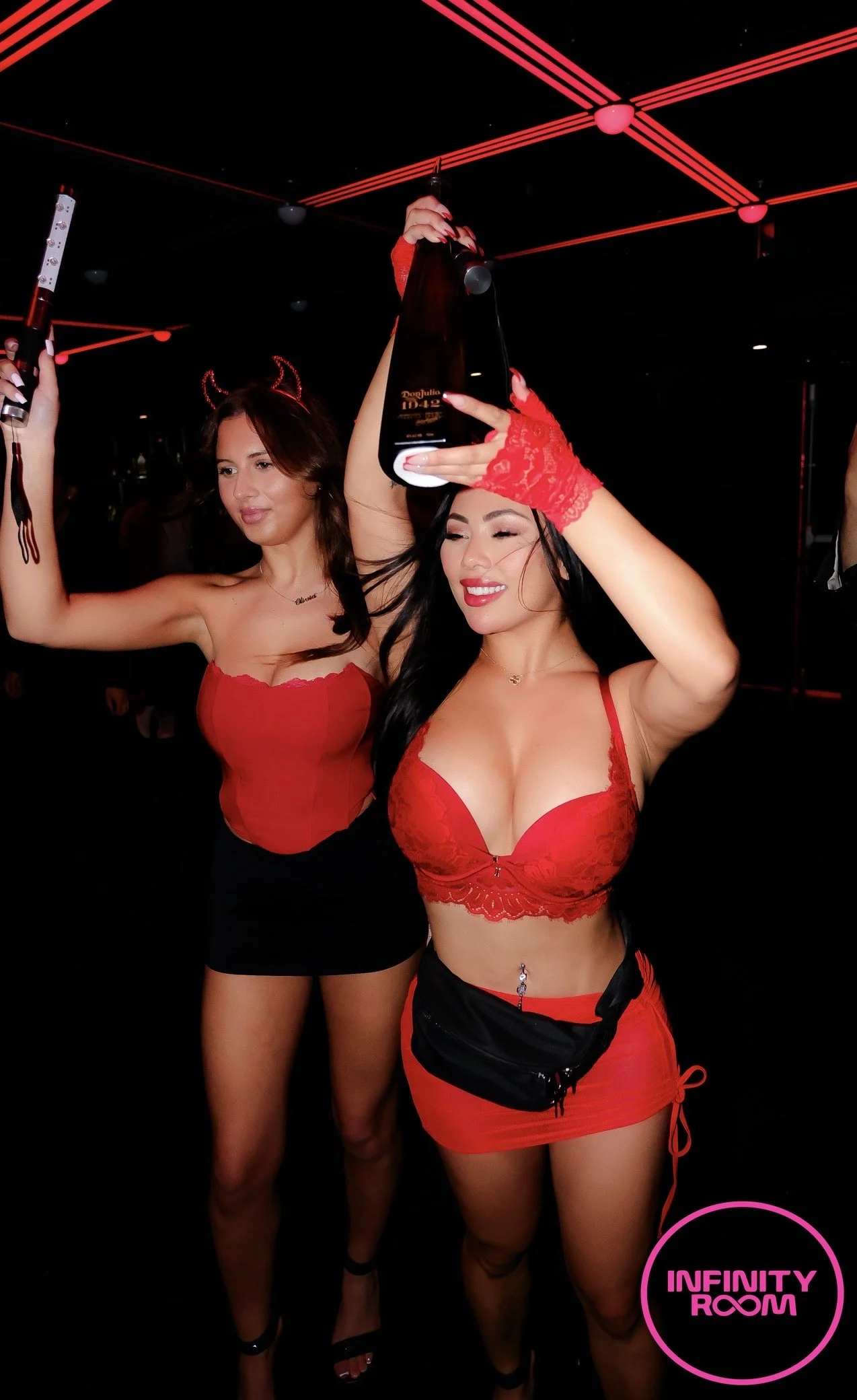 Two women in red outfits celebrating at a club with black background and red neon lights, one holding a bottle and the other a microphone, with a pink logo reading 'Infinity Room' in the corner.