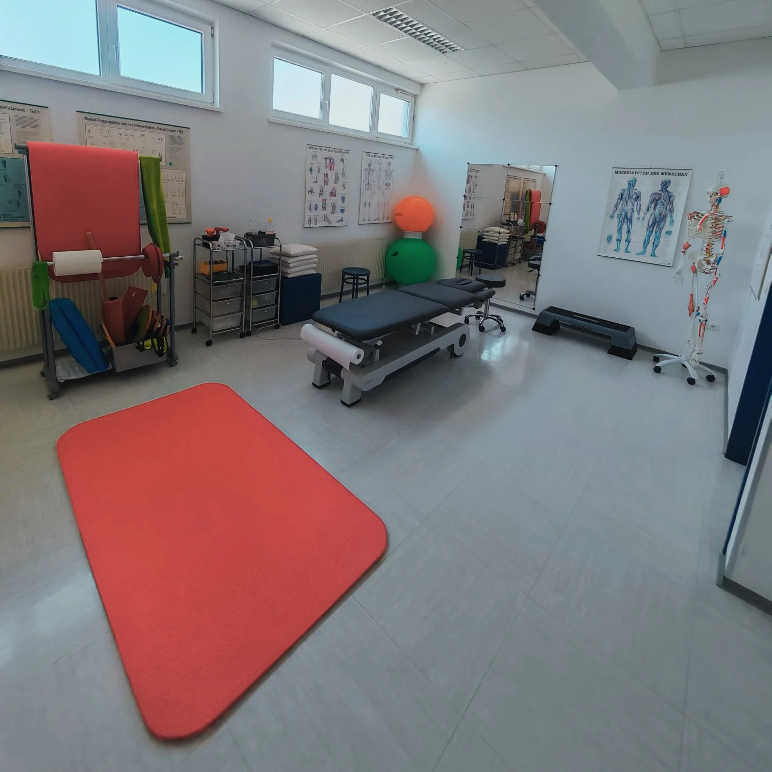 Therapieraum mit Turnmatte, Behandlungsbett, beweglichen medizinischen Geräten, Übungsmaterialien und anatomischen Postern an den Wänden.
