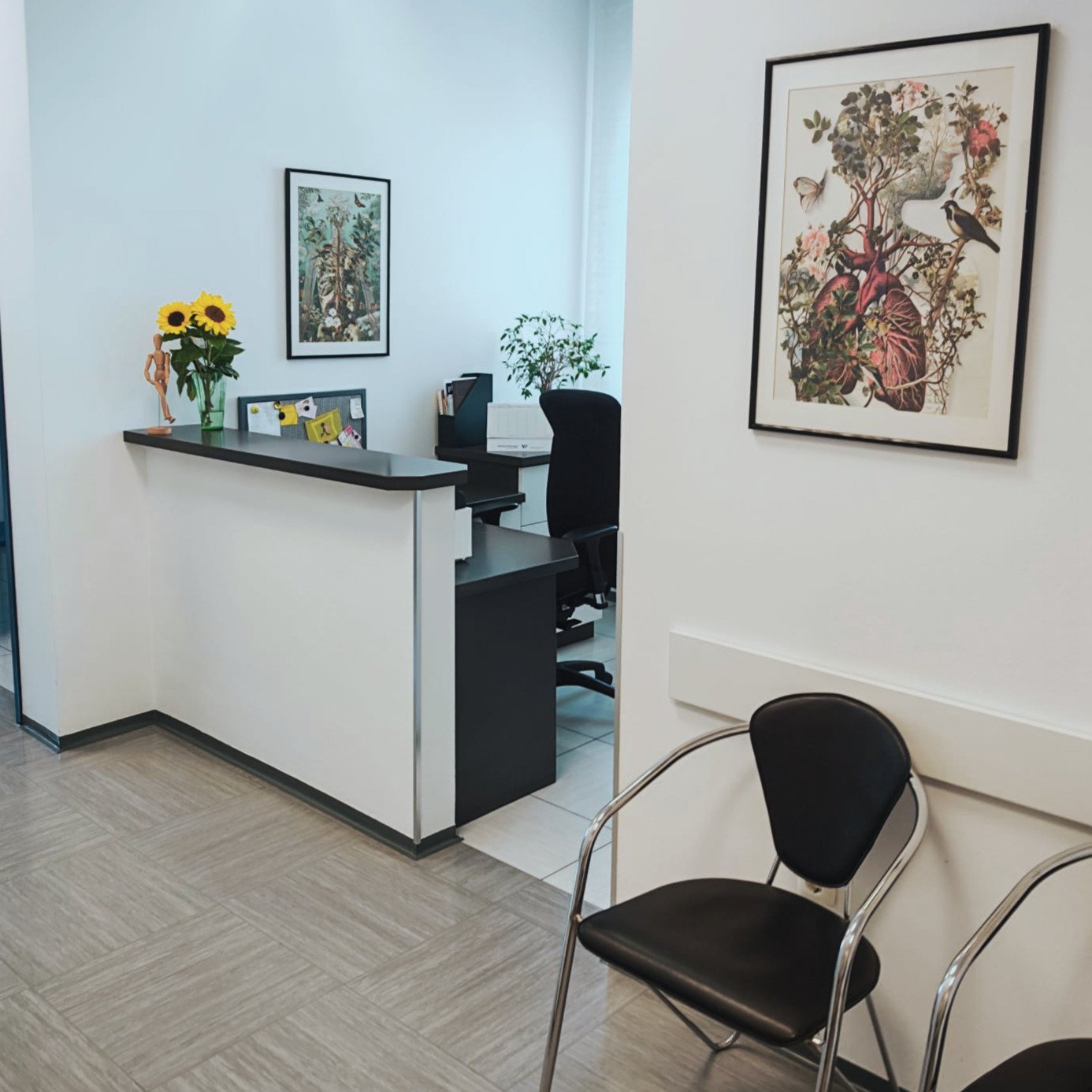 Wartezimmer mit Stühlen, einem Empfangstresen mit Sonnenblumen, verschiedenen Bildern an der Wand und einem Schreibtisch mit Stuhl im Hintergrund.