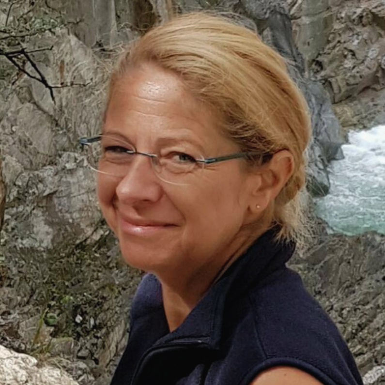 Eine lächelnde Frau mit roten Haaren und Brille steht vor einer felsigen Landschaft mit Fluss im Hintergrund.