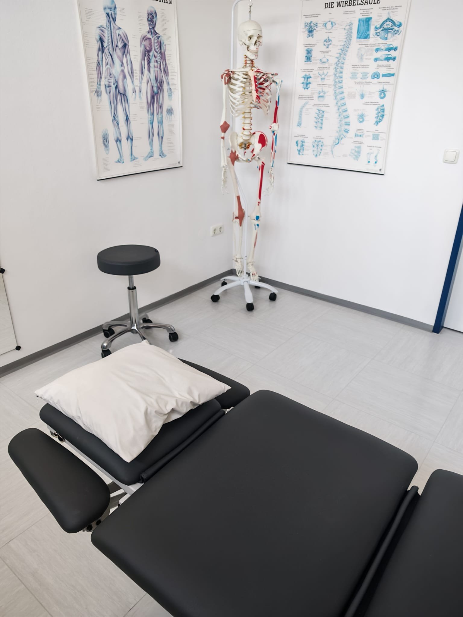 Ein Untersuchungstisch mit schwarzer Polsterung und verstellbaren Beinen, darauf liegt ein Kissen. Im Hintergrund gibt es ein Knochenmodell und anatomische Poster an der Wand in einem medizinischen Behandlungsraum.