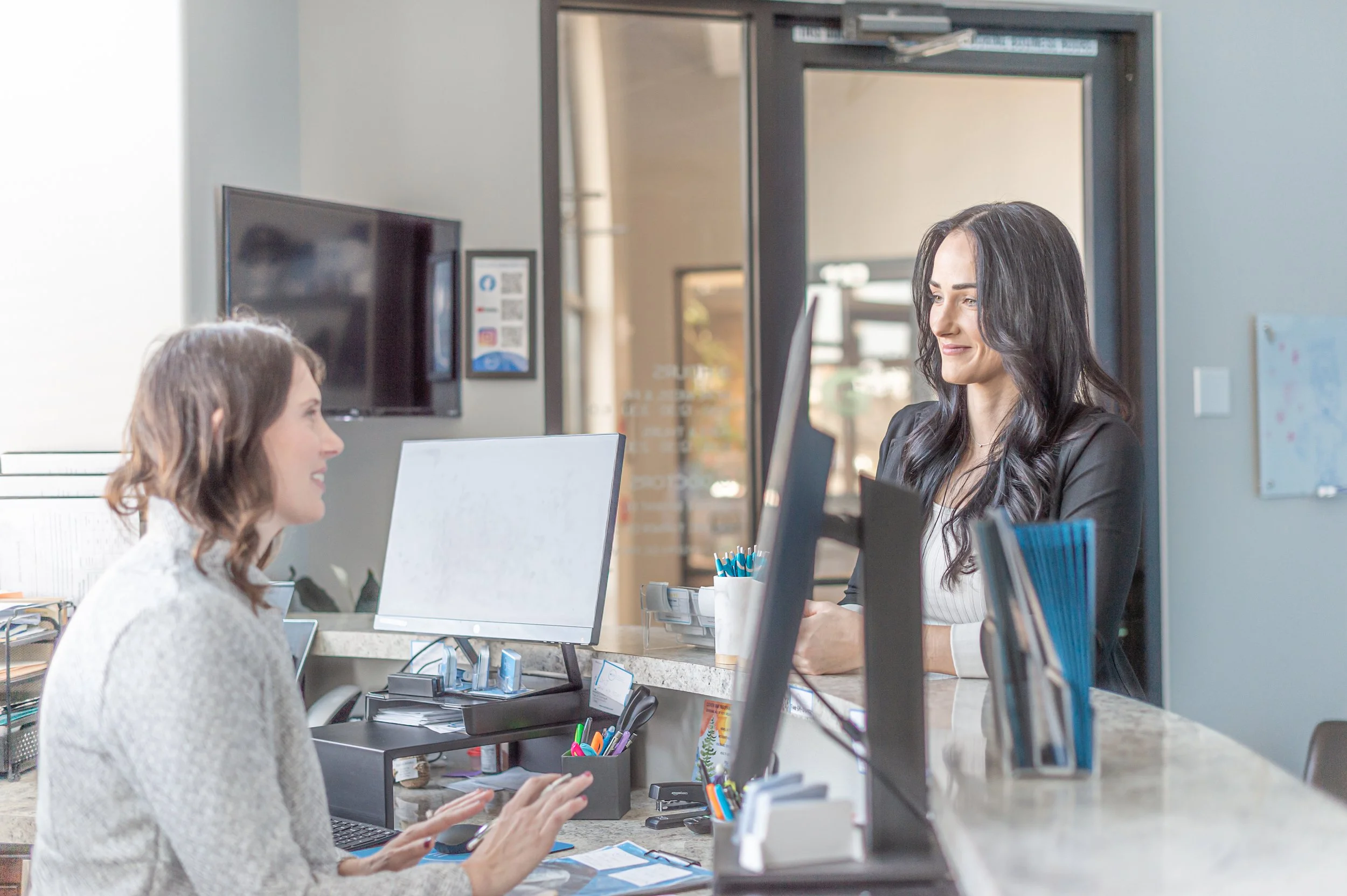 Two women at a reception desk, one sitting and one standing, engaging in conversation in an office environment with computers and office supplies.