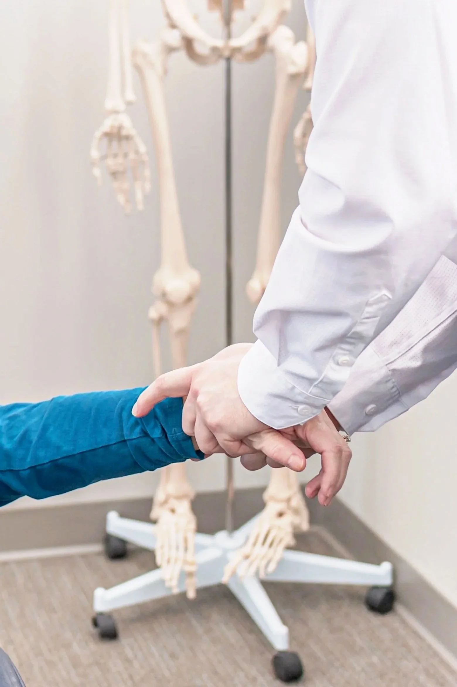 A healthcare professional holds a patient's hand in front of a skeleton model in a medical office.