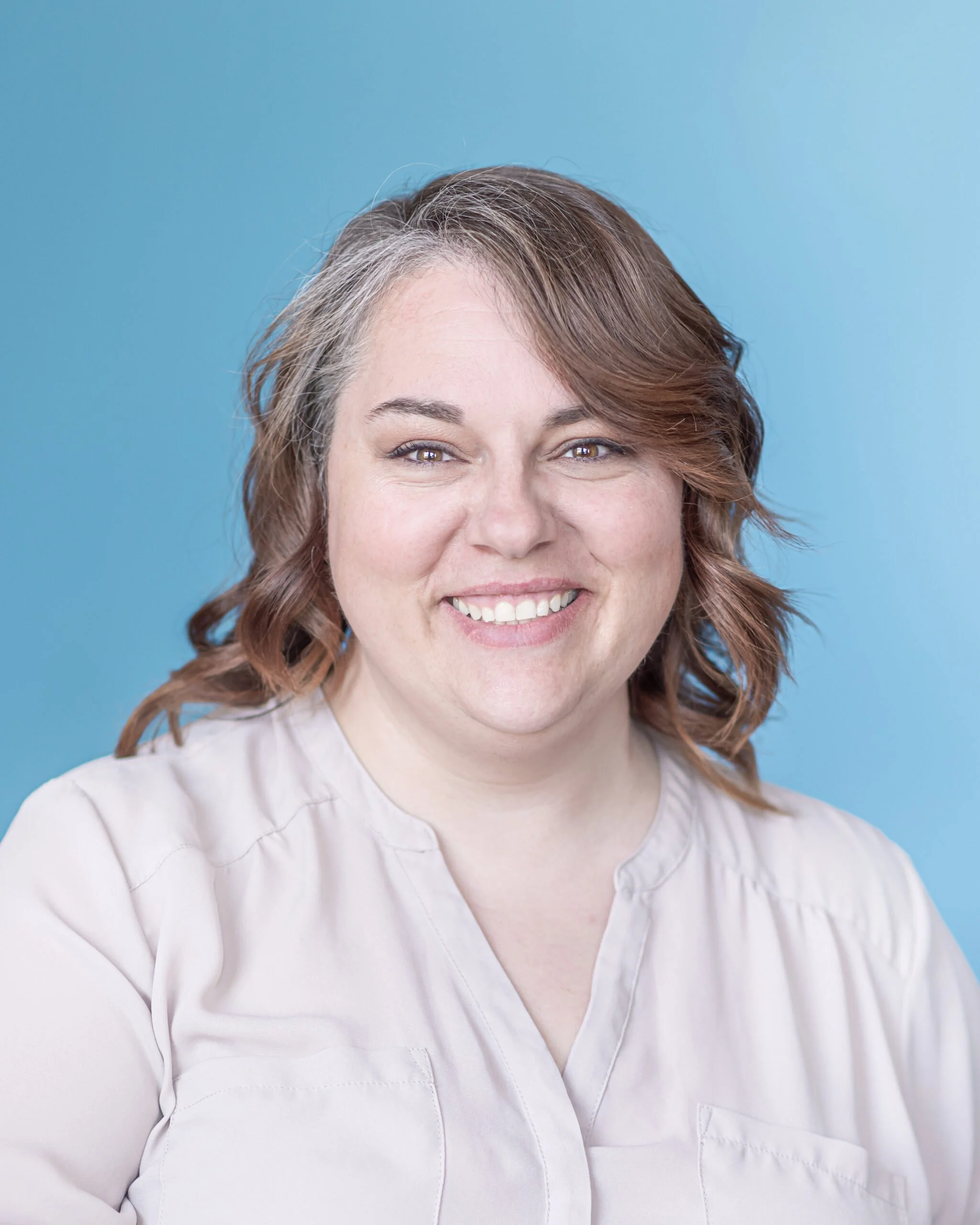 Close-up of a middle-aged woman with shoulder-length wavy brown hair with gray streaks, smiling against a solid light blue background, wearing a off-white blouse.